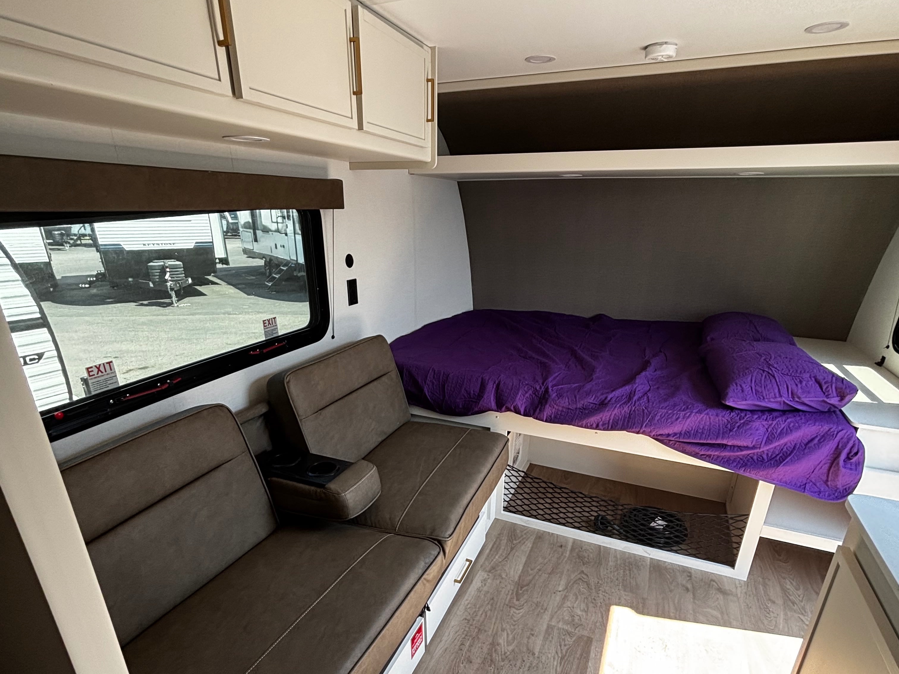 RV rental interior: A cozy space with a purple-sheeted bed, brown sofa with cupholders, overhead cabinets, and a window view.. Grand Design Transcend One 2027