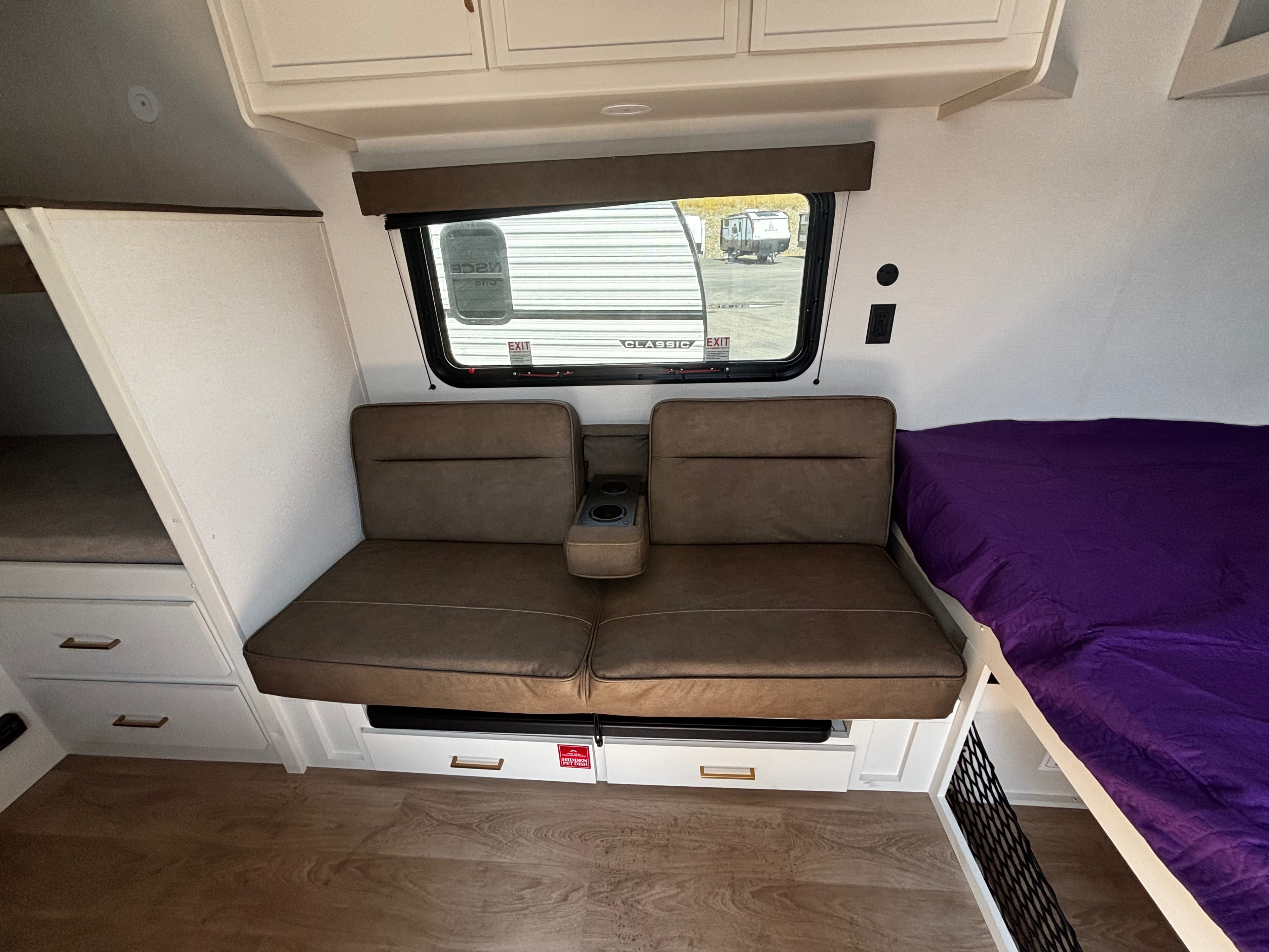 Bright RV rental interior: brown sofa with cupholders, window view, bunk beds, and a purple-sheeted bed.. Grand Design Transcend One 2027