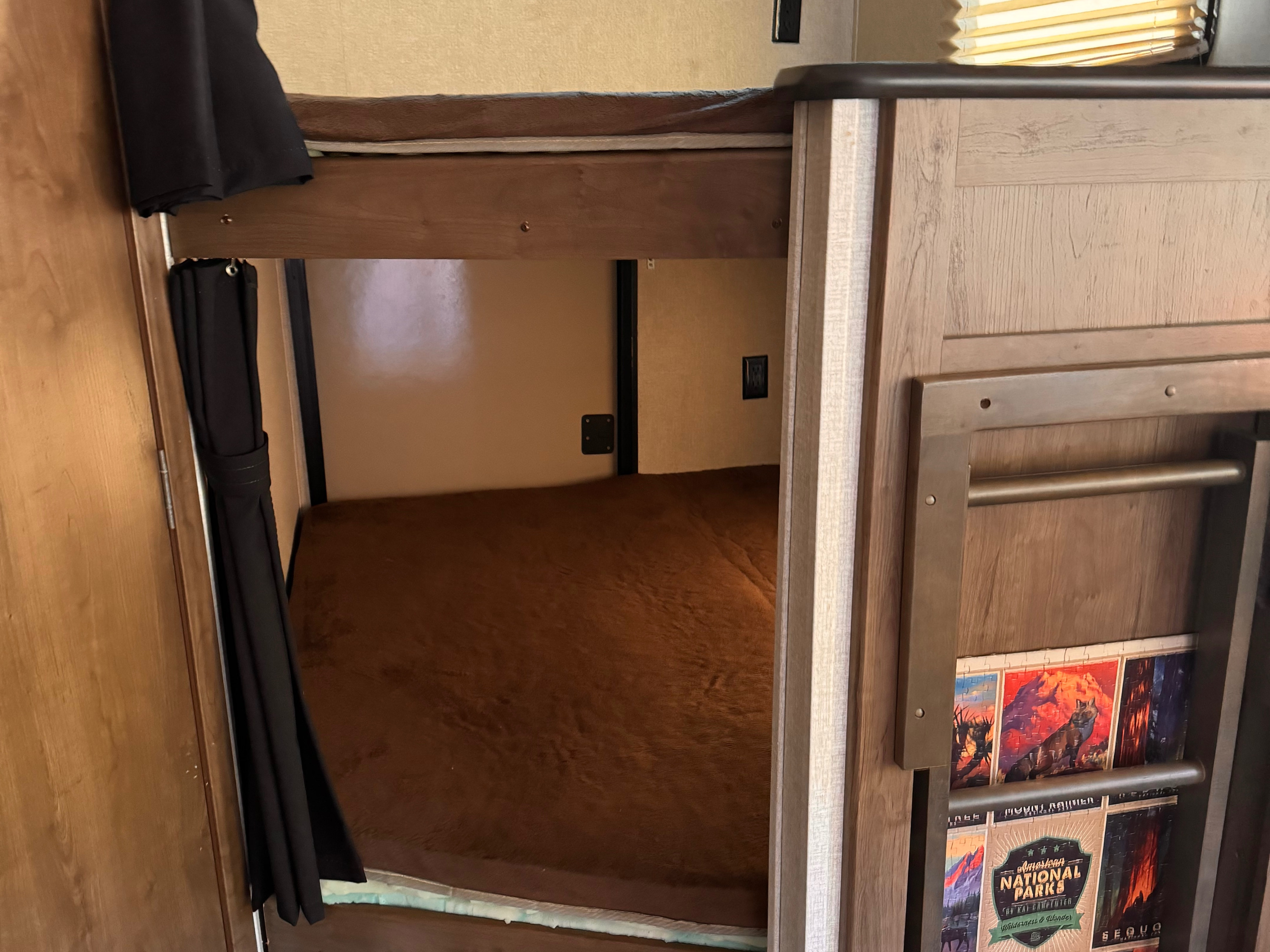 An RV rental features cozy bunk beds. The lower bunk has privacy curtains and a soft brown blanket, with National Parks art.. Keystone RV Cougar 2019