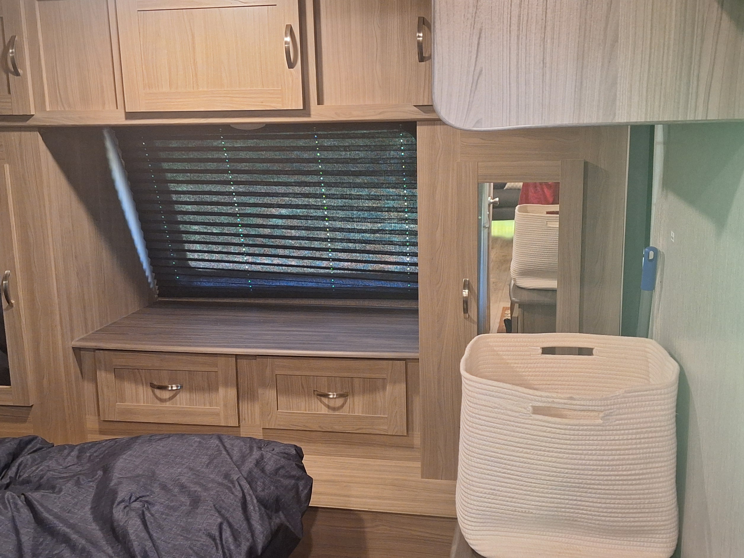 RV rental bedroom with light wood storage cabinets, window blinds, a bed, reflected mirror view, and a white rope basket.. Coachmen Liberty express 2021