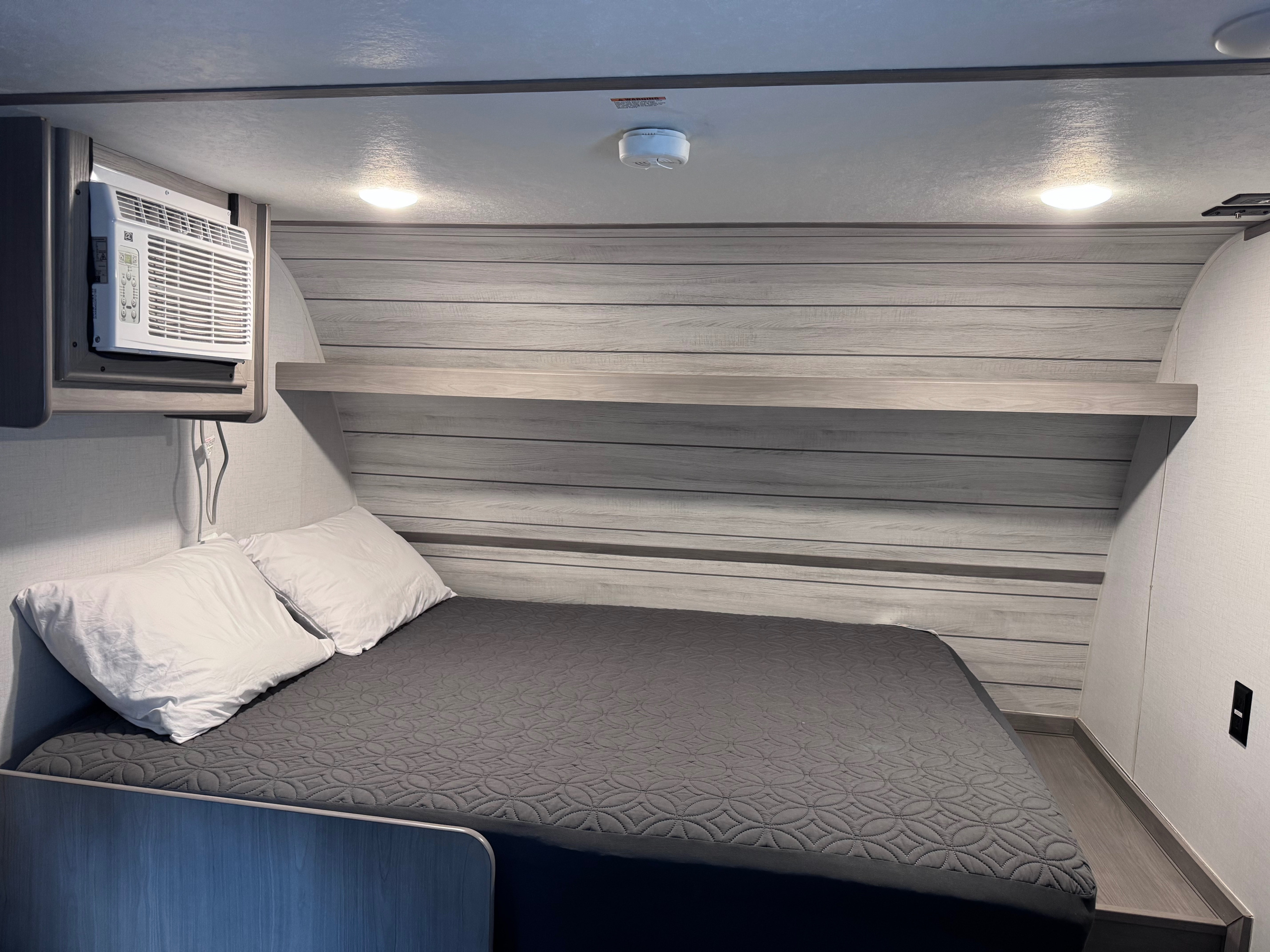RV rental bedroom: compact space with a cozy bed, white pillows, AC unit, wood-paneled wall, ceiling lights & smoke alarm.. Dutchmen Coleman 2024