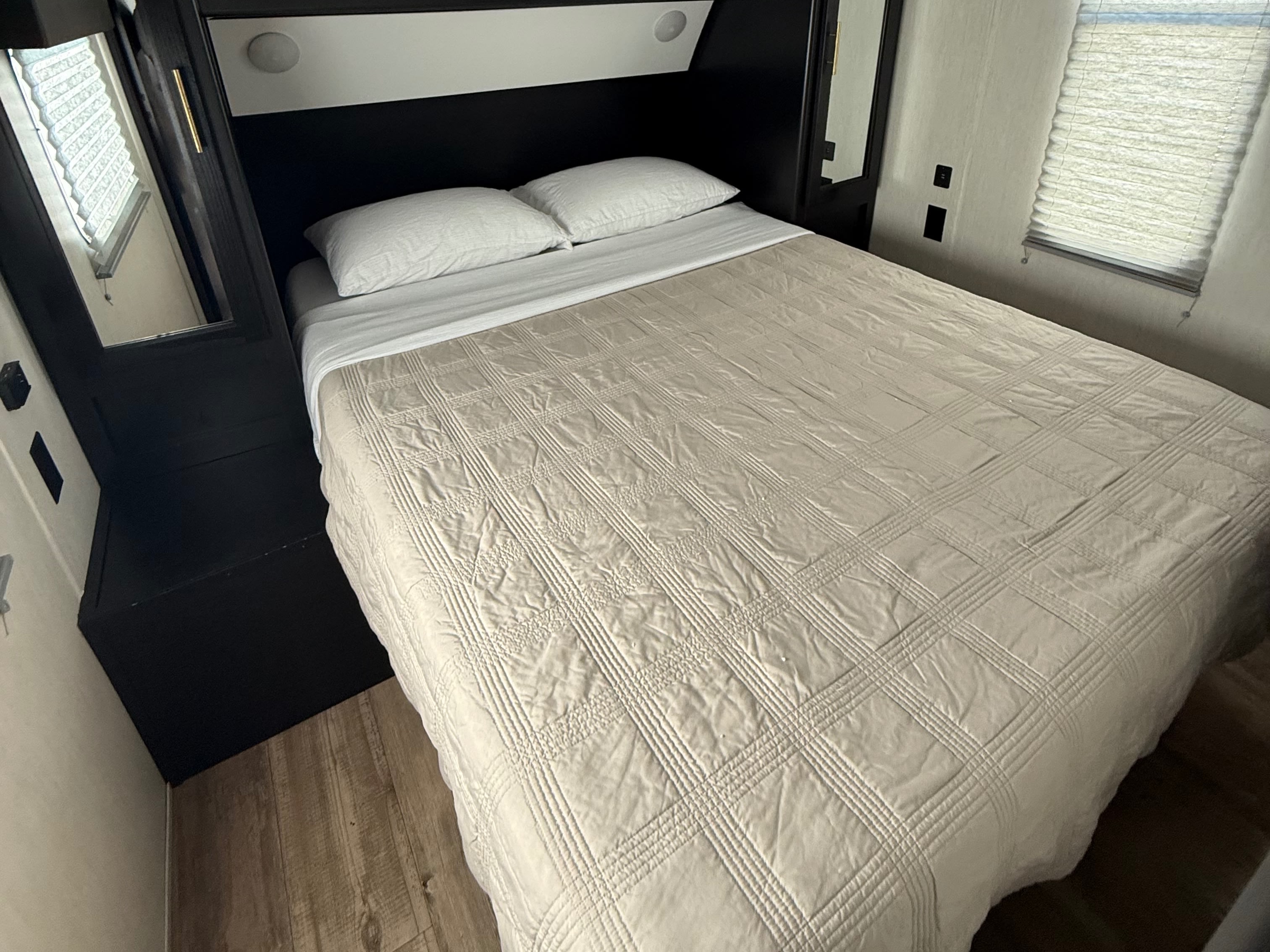 Cozy RV rental bedroom features a bed with white pillows, a beige quilted comforter, a mirror, and a window with blinds.. Forest River Cherokee Grey Wolf 2022