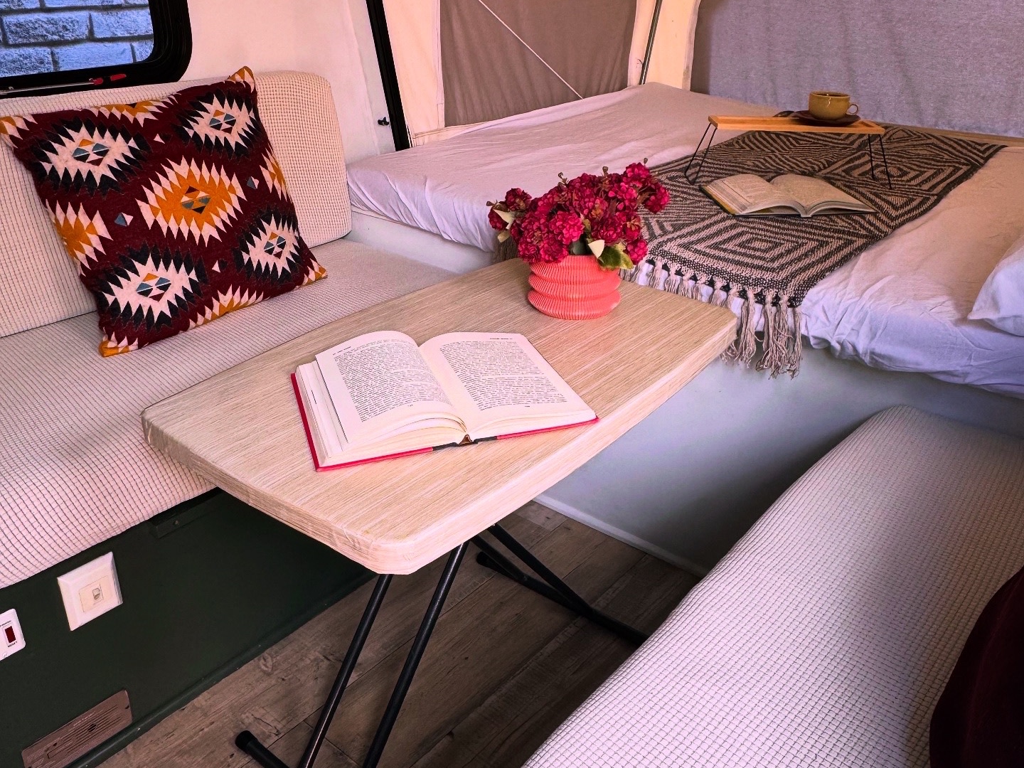 Cozy pop-up RV rental interior featuring a patterned pillow, a table with books & flowers, and a bed with a blanket. Ideal for reading.. Sportsmen Classic 2014