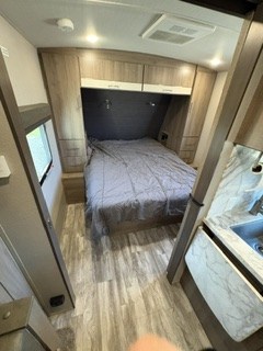 RV rental interior: A modern bedroom with a grey bed, overhead cabinets, wood-look floor, and a partial kitchen view.. Grand Design Imagine 2023