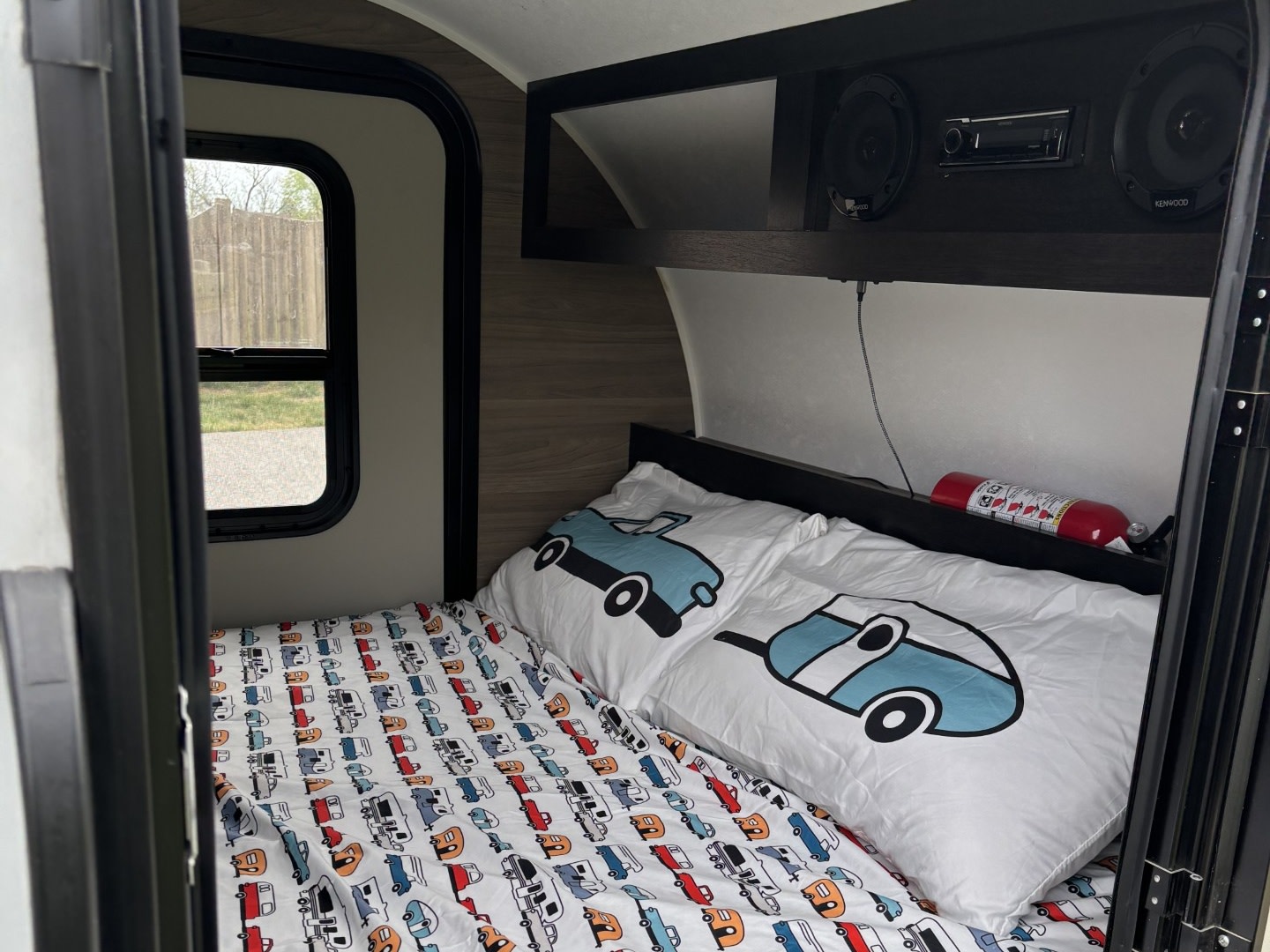 Cozy rv rental interior with camper-themed bedding on a bed, a window, overhead stereo system, and a red fire extinguisher.. Braxton Creek Bushwhacker 2024