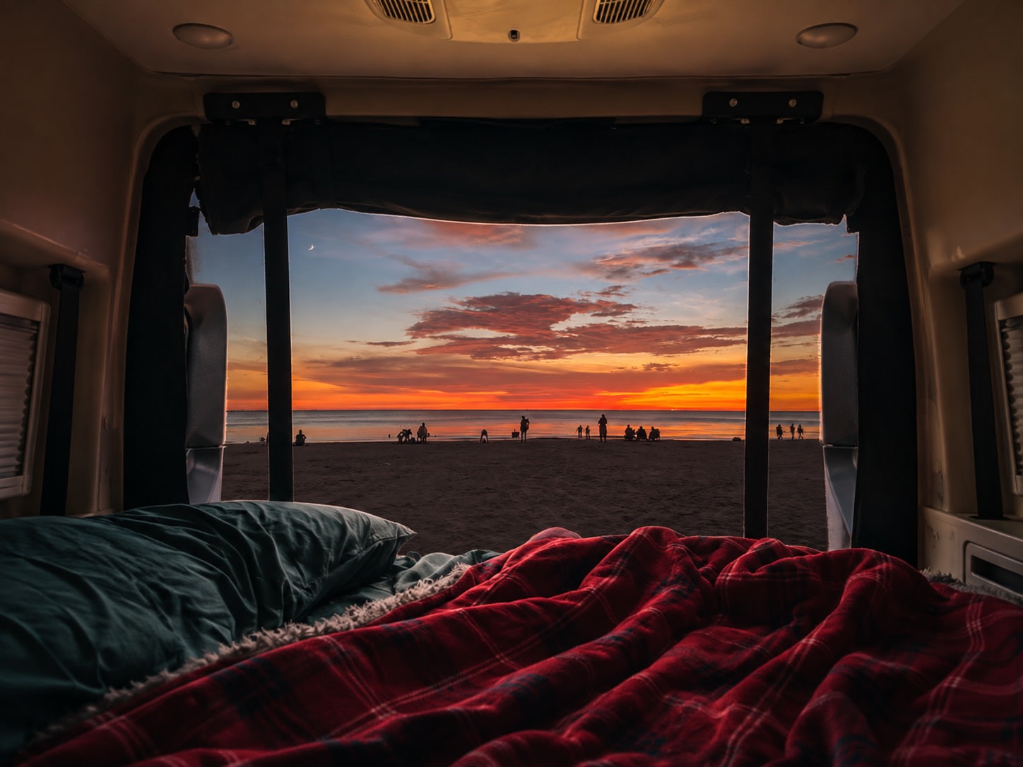 Cozy RV rental bed overlooking a vibrant beach sunset with people, ocean, and a crescent moon. Perfect travel view.. Entegra Coach Launch 2024