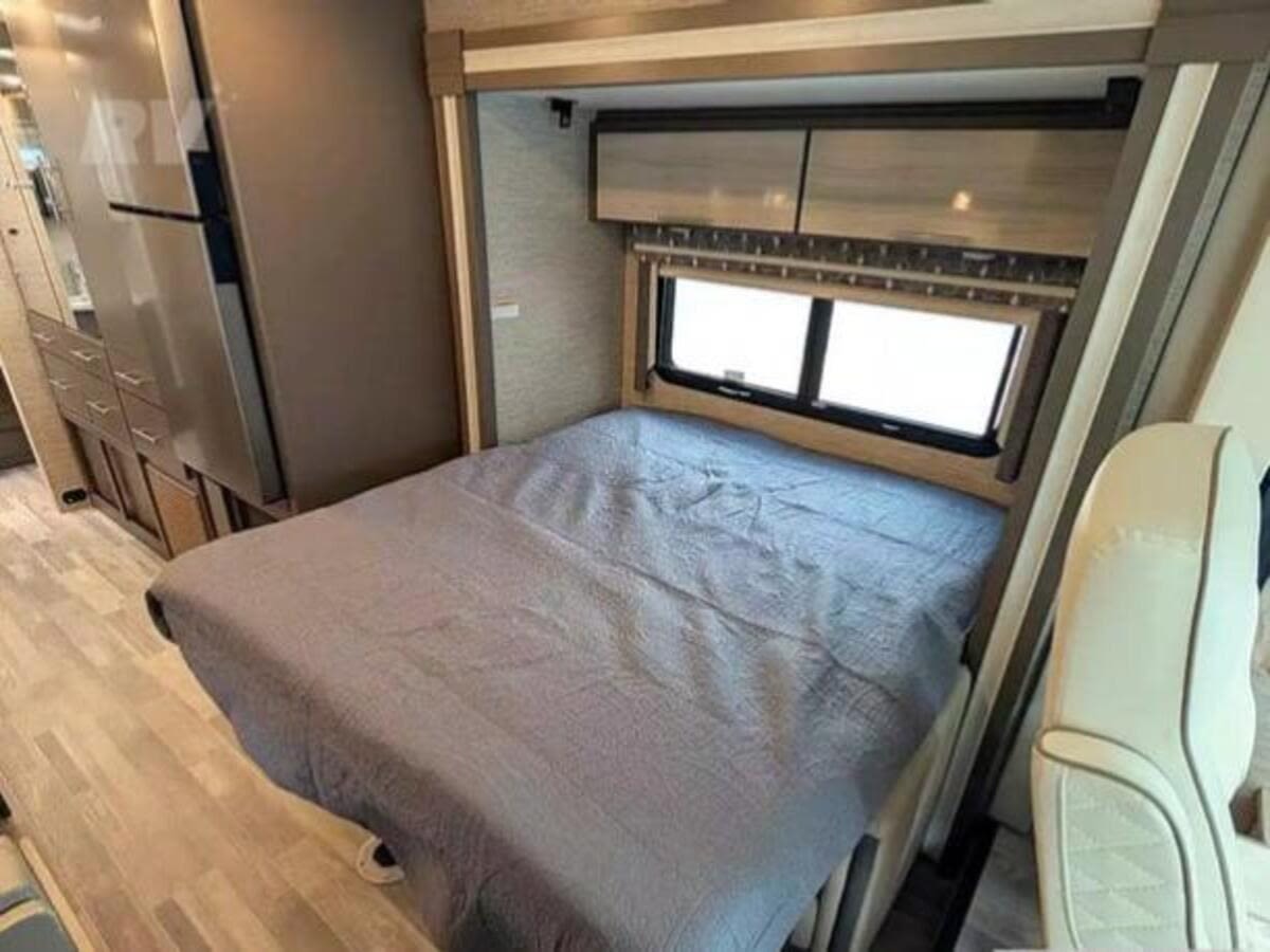 Comfortable RV rental interior: A made bed with grey bedding, a window, overhead cabinets, and a fridge on the left.. Thor Axis 2023