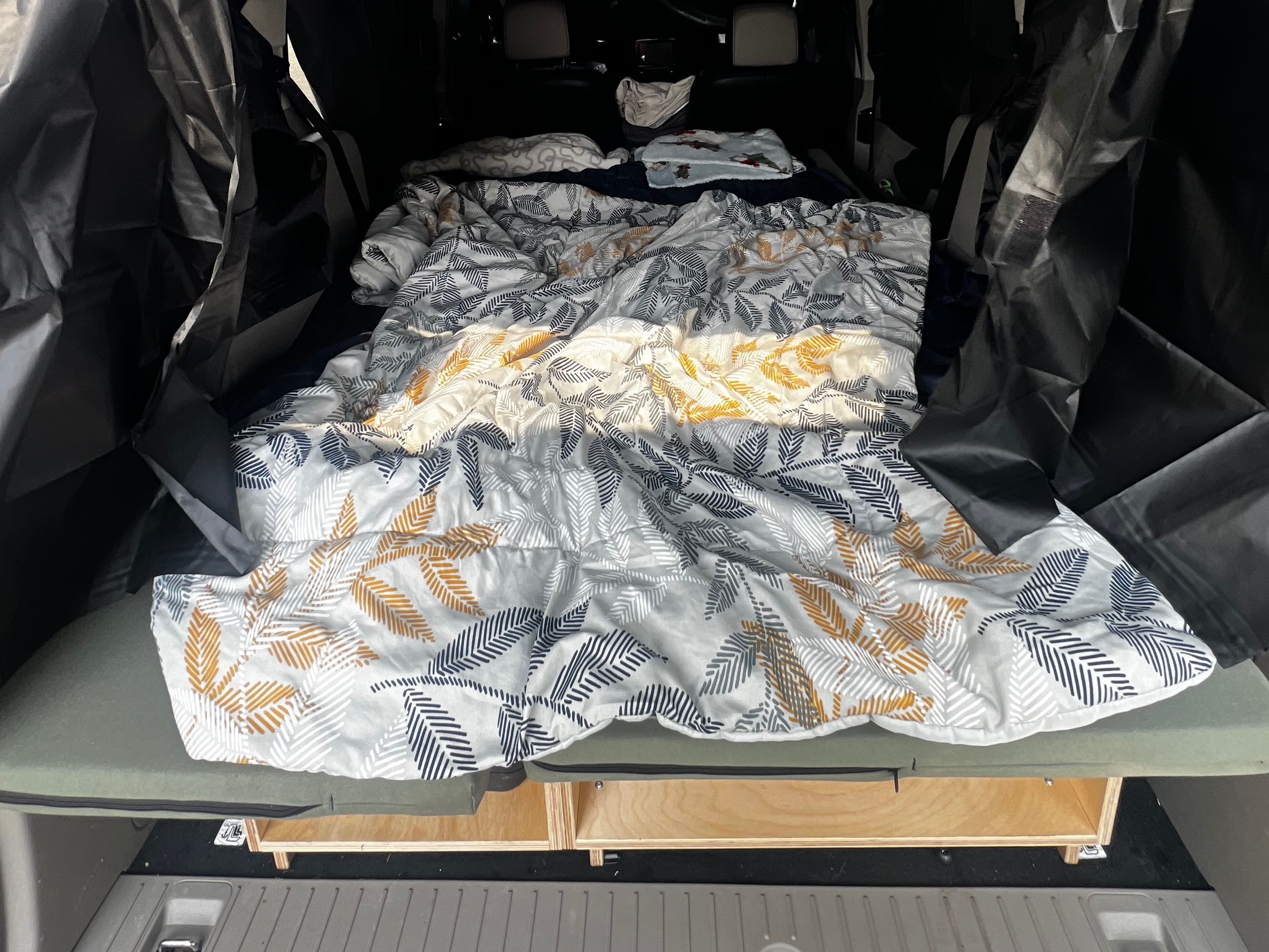 RV rental car camping setup: a cozy bed with gray, gold, and navy leaf-patterned comforter over wooden storage.. Ford transit 2019