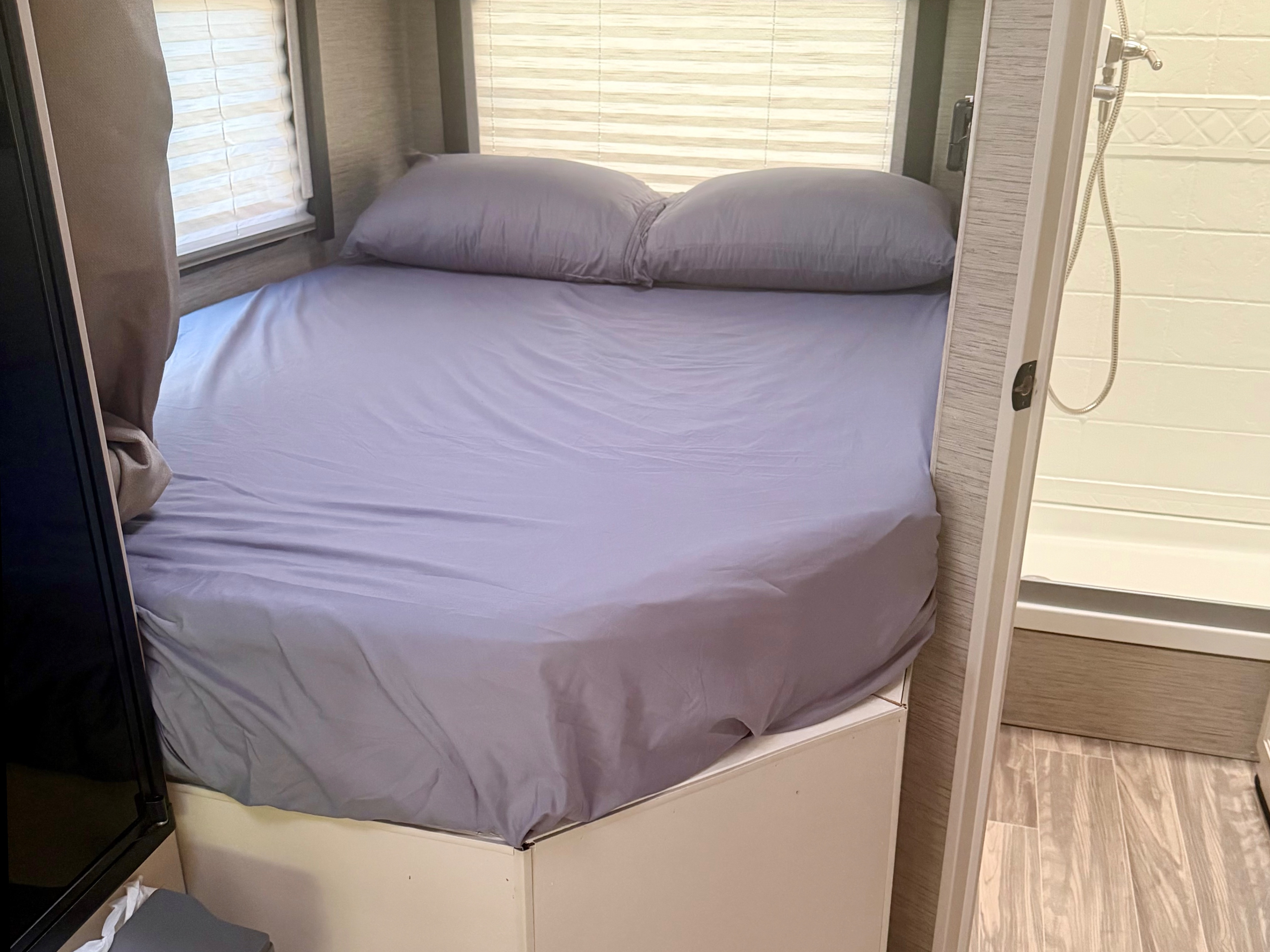 An RV rental bedroom with a neatly made bed, purple sheets, window blinds, and a glimpse of the shower area.. Thor Four Winds 2023