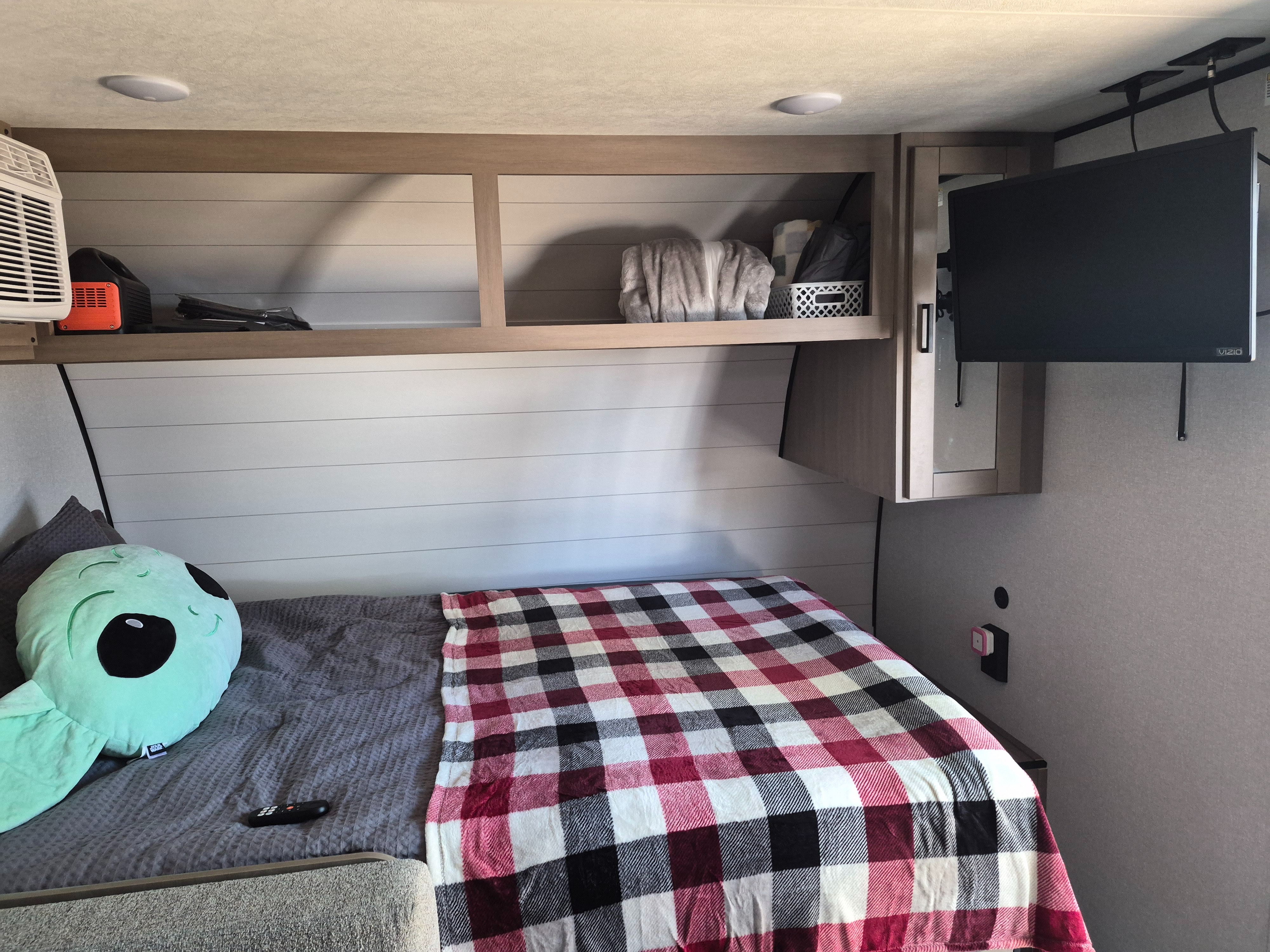 Cozy RV rental bedroom with a plaid-covered bed, alien pillow, overhead storage shelves, and a wall-mounted TV.. Open Range Colt 17BH 2026