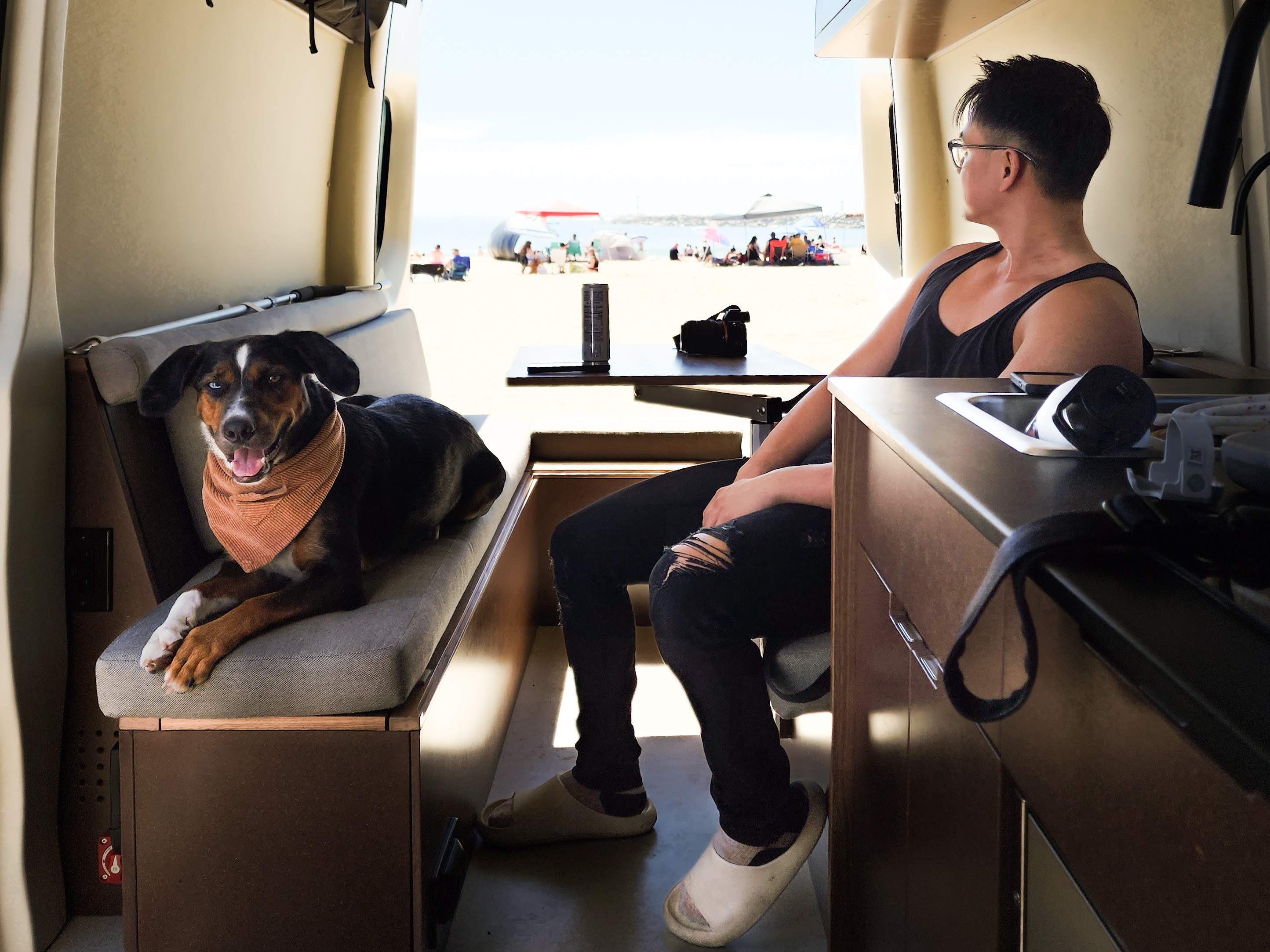 Inside an RV rental: A man and dog enjoy beach views. Overhead storage & kitchen are visible, with people on the sand outside.. Mercedes Sprinter 2021