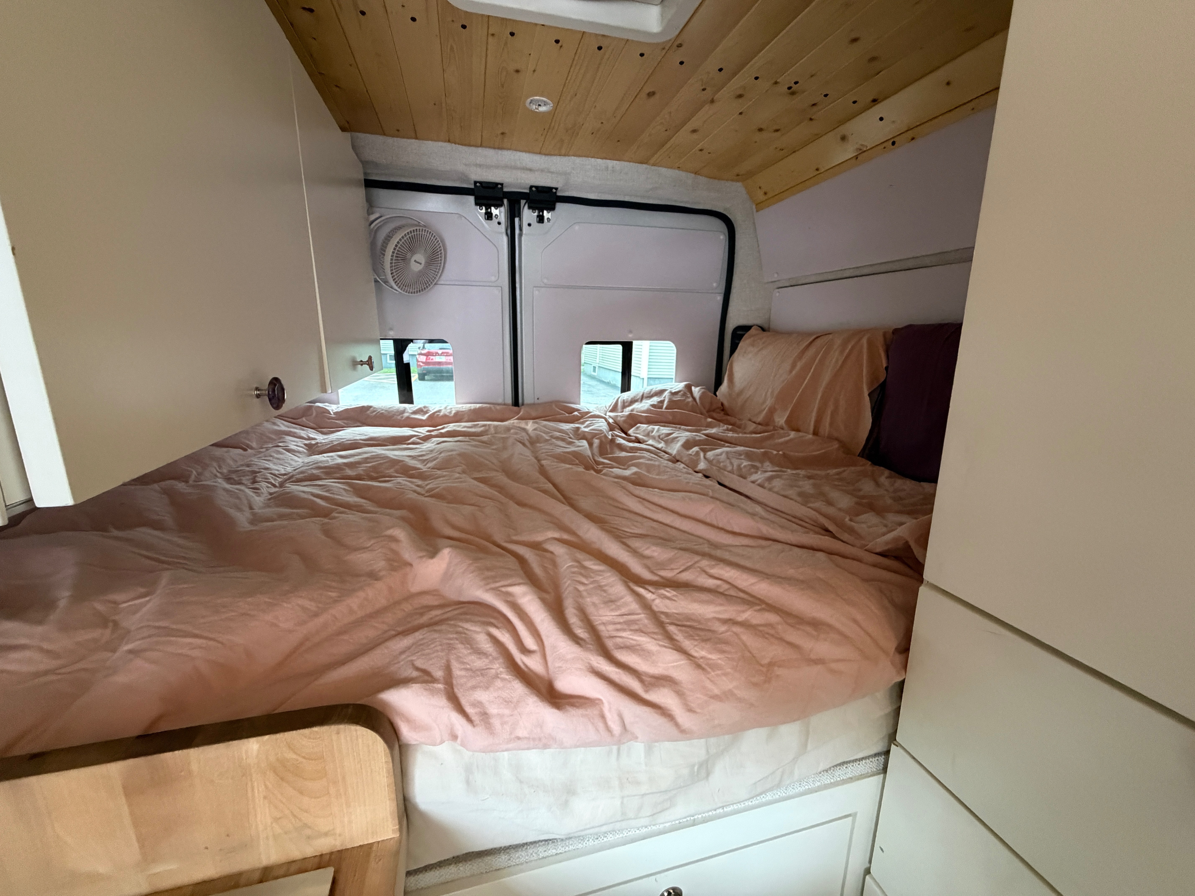 Cozy RV rental interior: a comfy bed with peach bedding, wooden ceiling, fan, and rear windows for a view.. Ram Promaster 2024