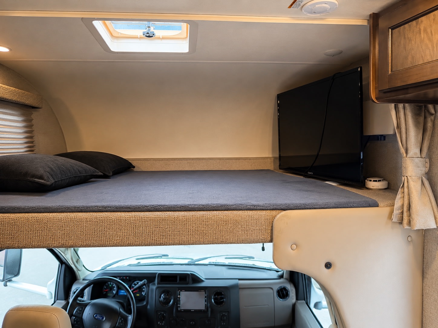 Inside an RV rental: a cab-over bunk features a bed, pillows, TV, and skylight. The vehicle's dashboard is below.. Thor Freedom Elite 2020