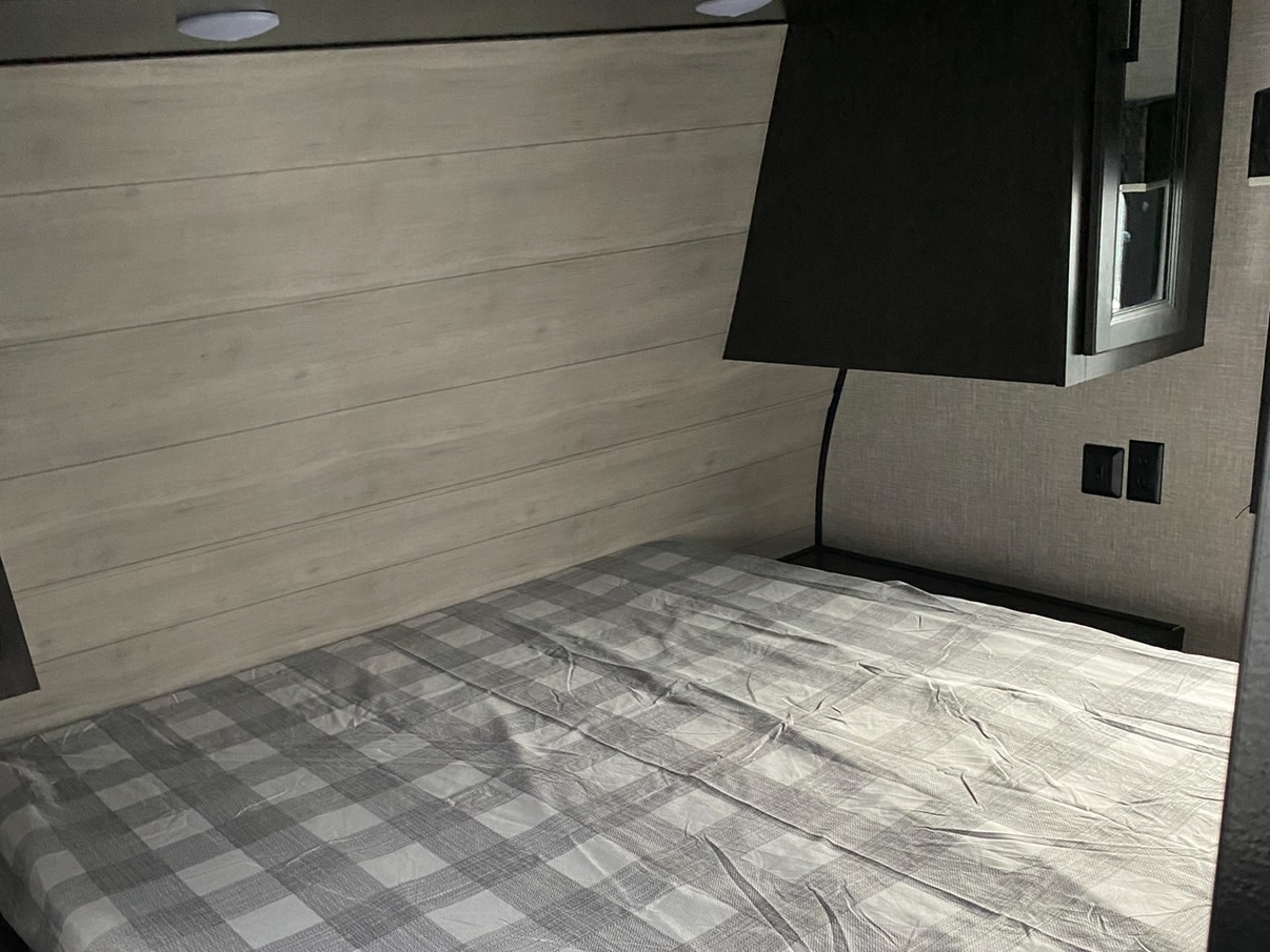 RV rental bedroom with a bed featuring gray plaid sheets, light wooden wall, and dark overhead cabinet.. Jayco Jay Flight SLX 2022