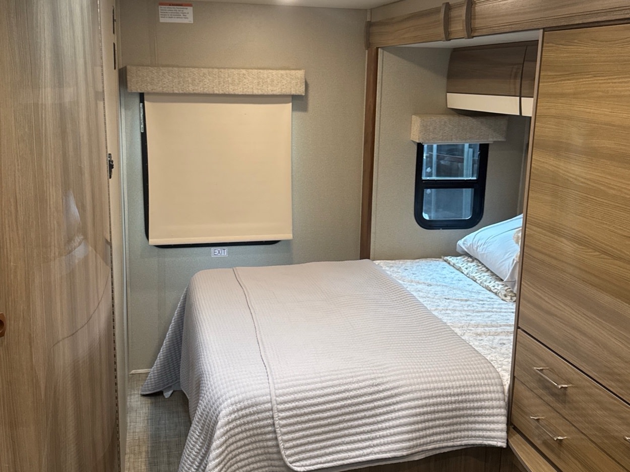 View inside an RV rental bedroom showing a bed, two windows (one with exit sign), and built-in wooden storage.. Entegra Coach Qwest 2018