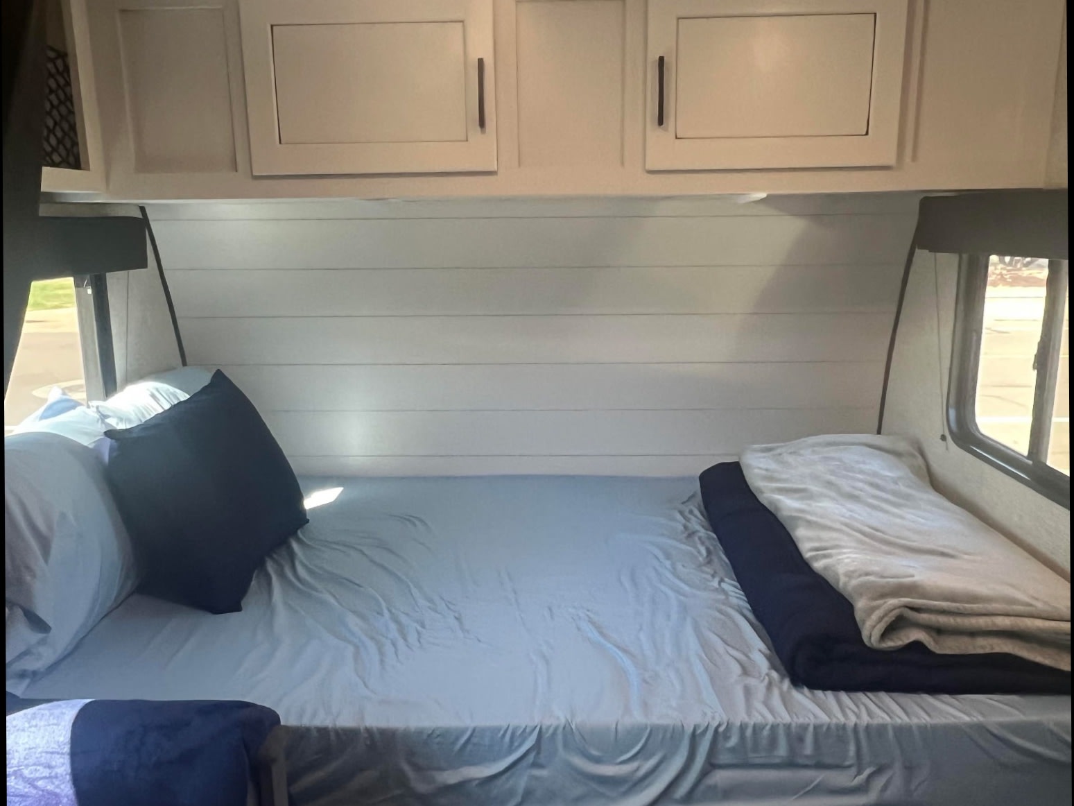 RV rental bedroom featuring a bed with light blue sheets, pillows, dark blankets, shiplap wall, and overhead cabinets.. Jayco Jay Flight SLX Baja Edition 2024