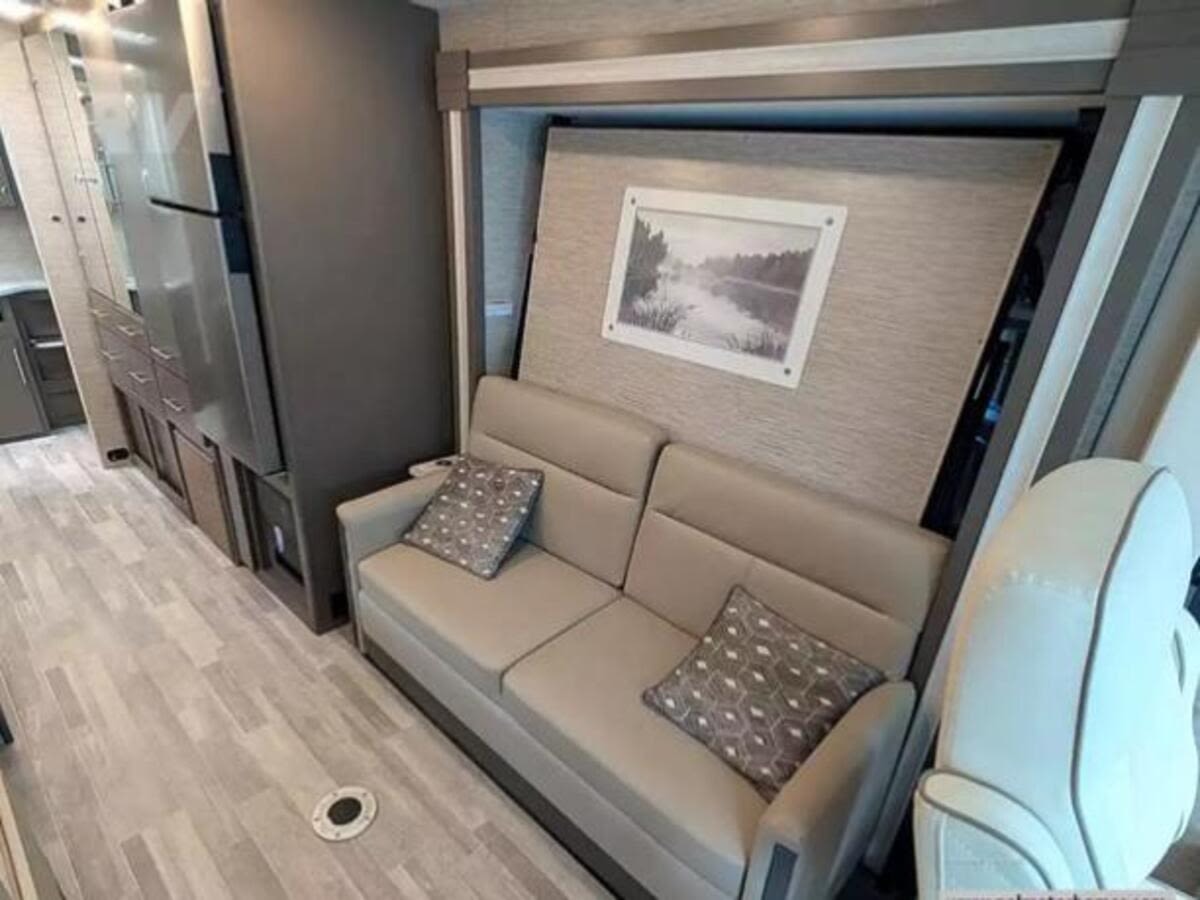 Modern RV rental interior featuring a beige sofa, kitchen with fridge, and a wall panel with art. Spacious motorhome living area.. Thor Axis 2023