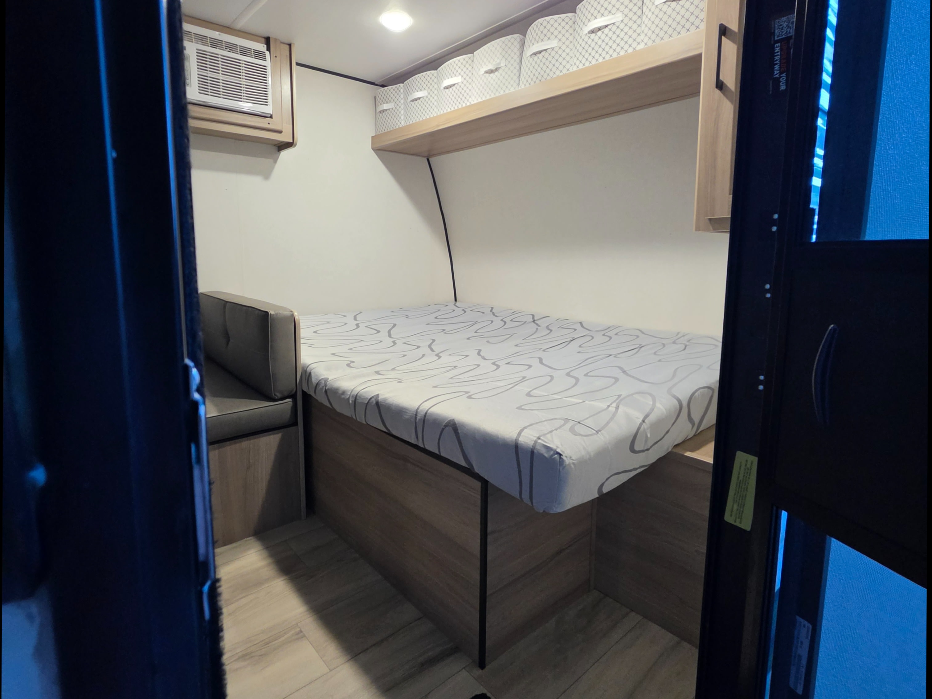 Cozy RV rental interior with a fitted bed, small sofa, overhead storage bins, and an AC unit for comfortable travel.. Keystone RV Coleman 2025