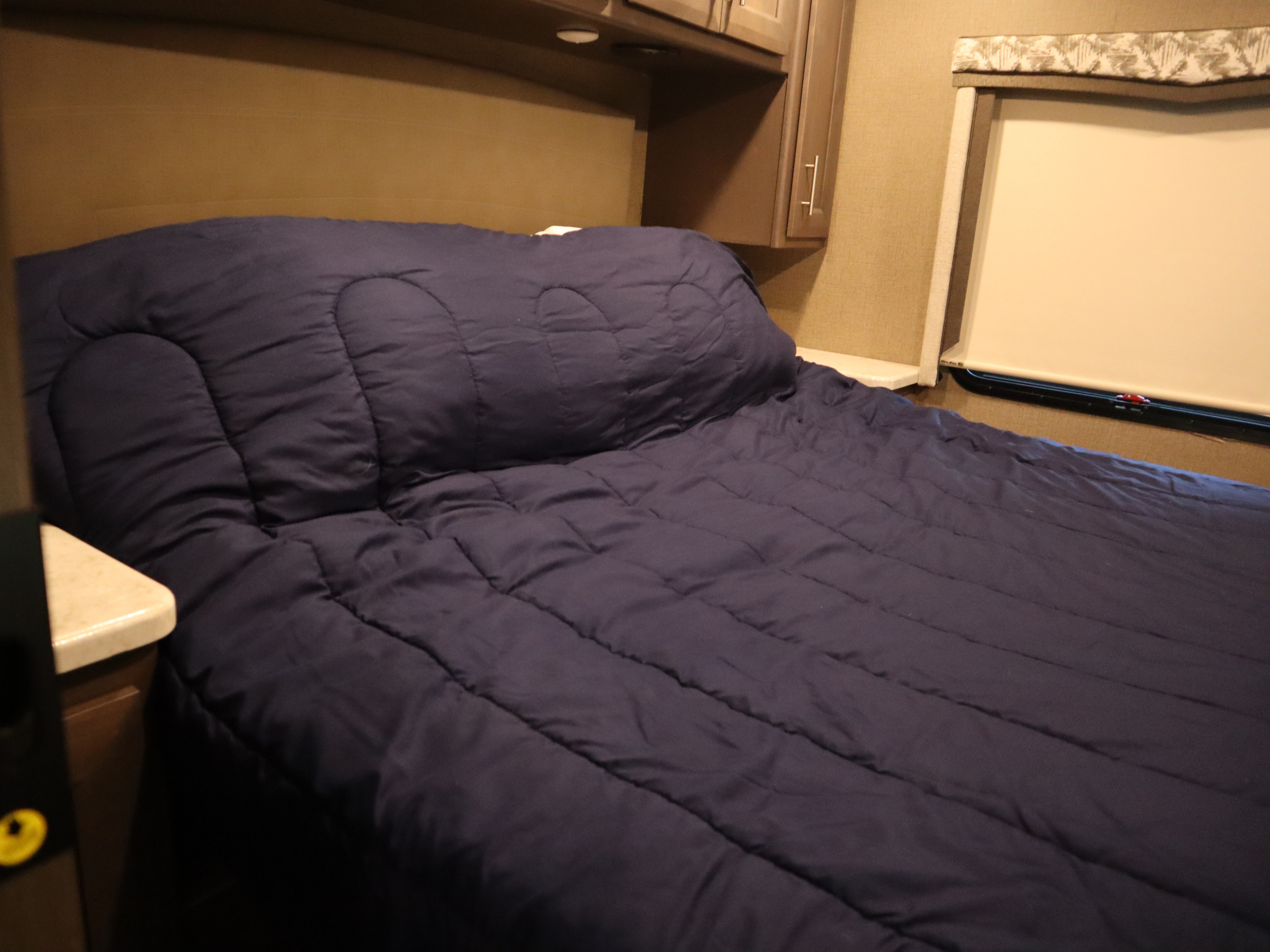 Cozy RV rental bedroom with a dark blue quilted comforter, overhead cabinets, and a window with a blind.. Thor Magnitude 2021