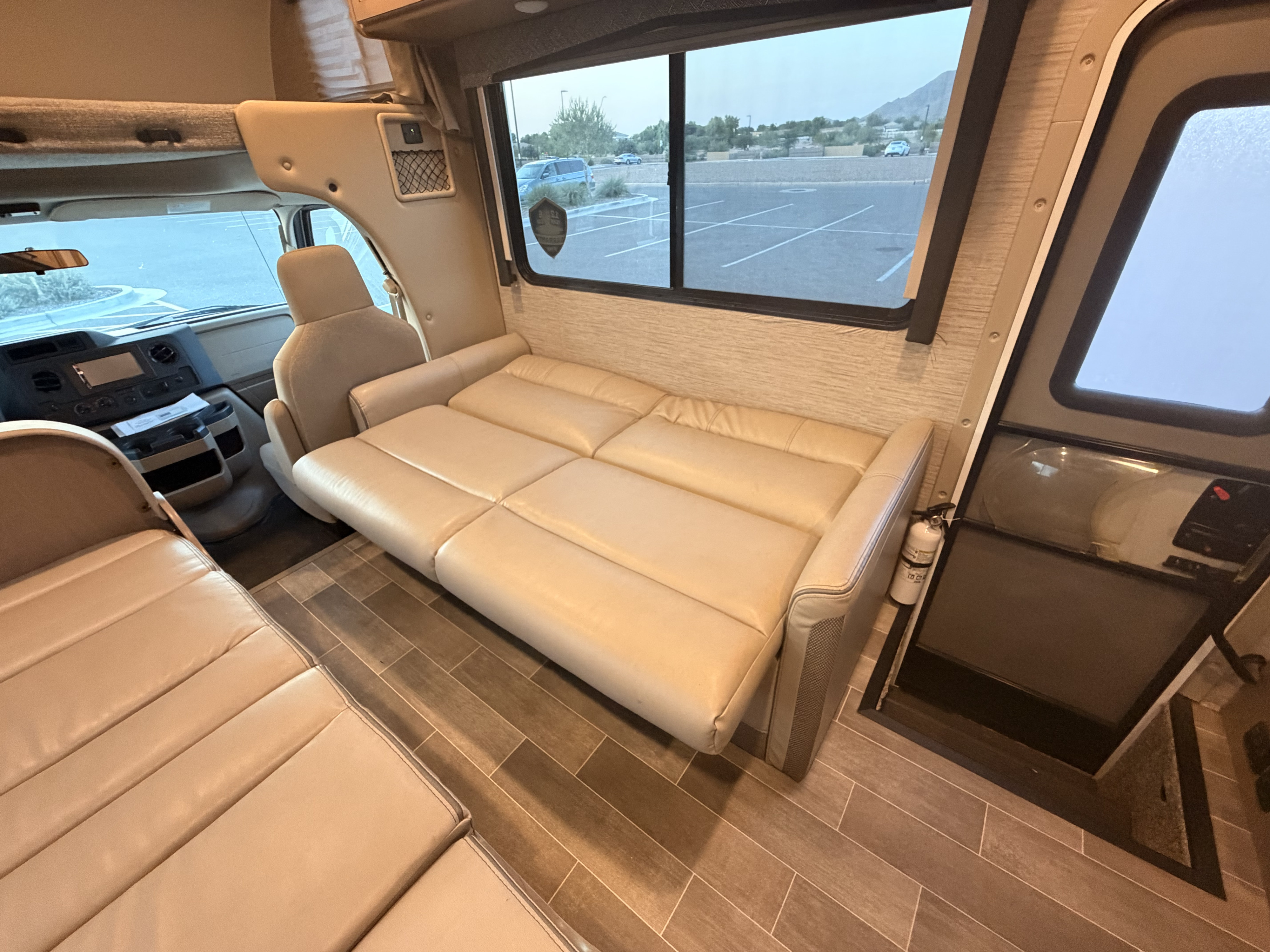 View inside an rv rental: A beige sofa-bed is pulled out, facing the driver's cabin. A window shows a parking lot and mountains outside.. Thor Chateau 2022