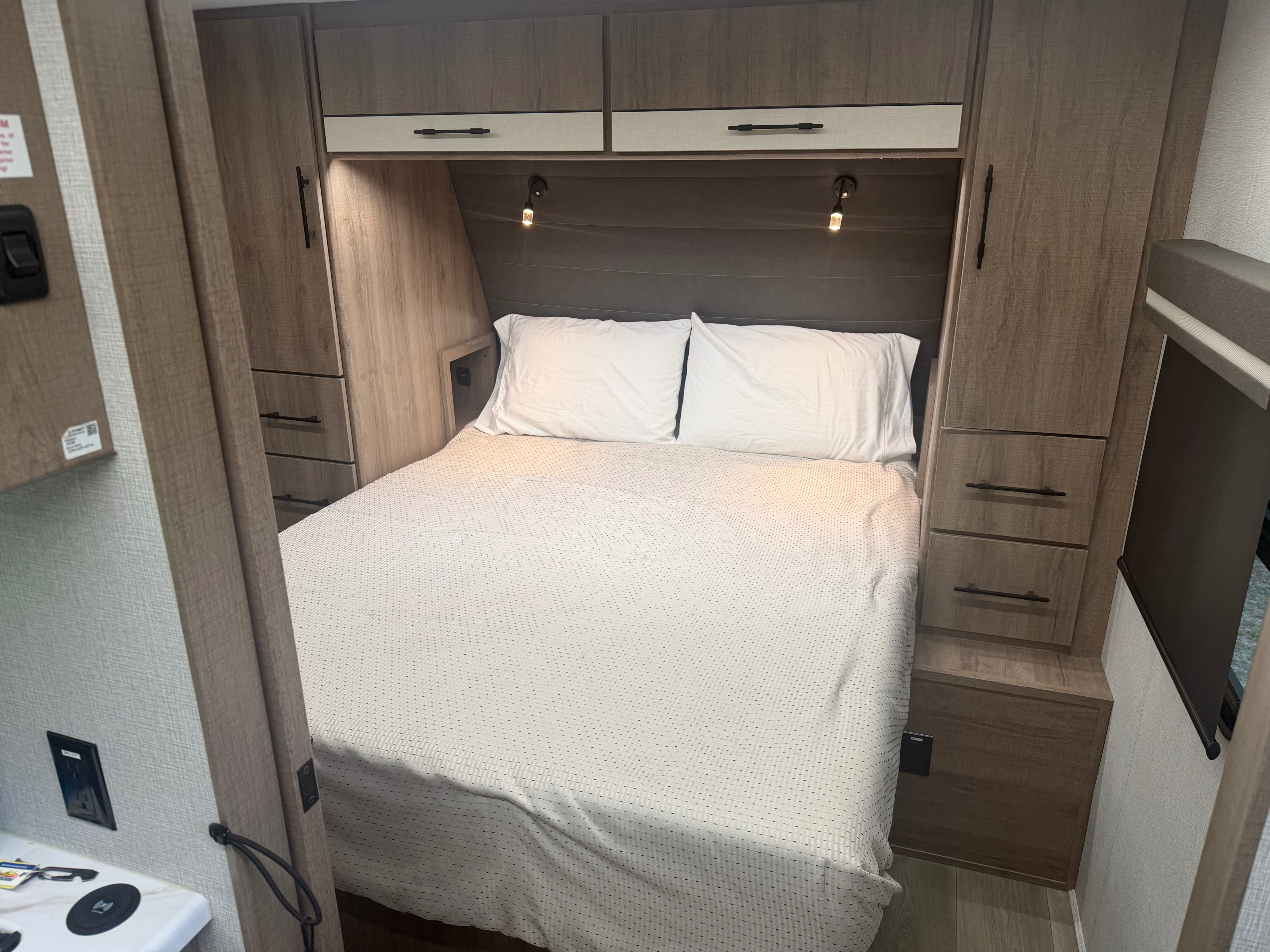 Cozy RV rental bedroom features a comfortable bed, overhead cabinets, and handy side storage with reading lights.. Grand Design Imagine XLS 2024