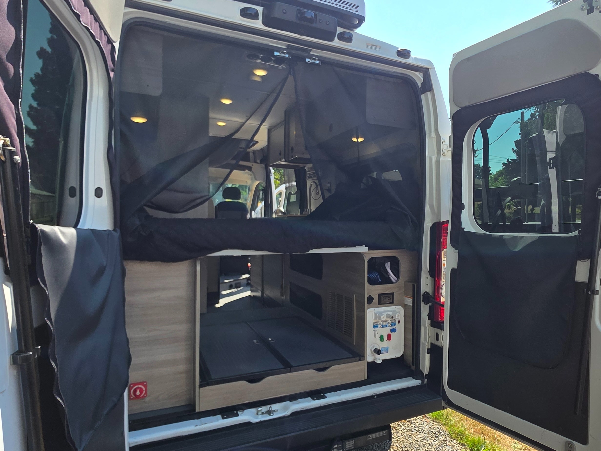 Open rear view of an RV rental van's organized interior: bed, mosquito net, storage, and external utility connections.. Winnebago Porto 2021