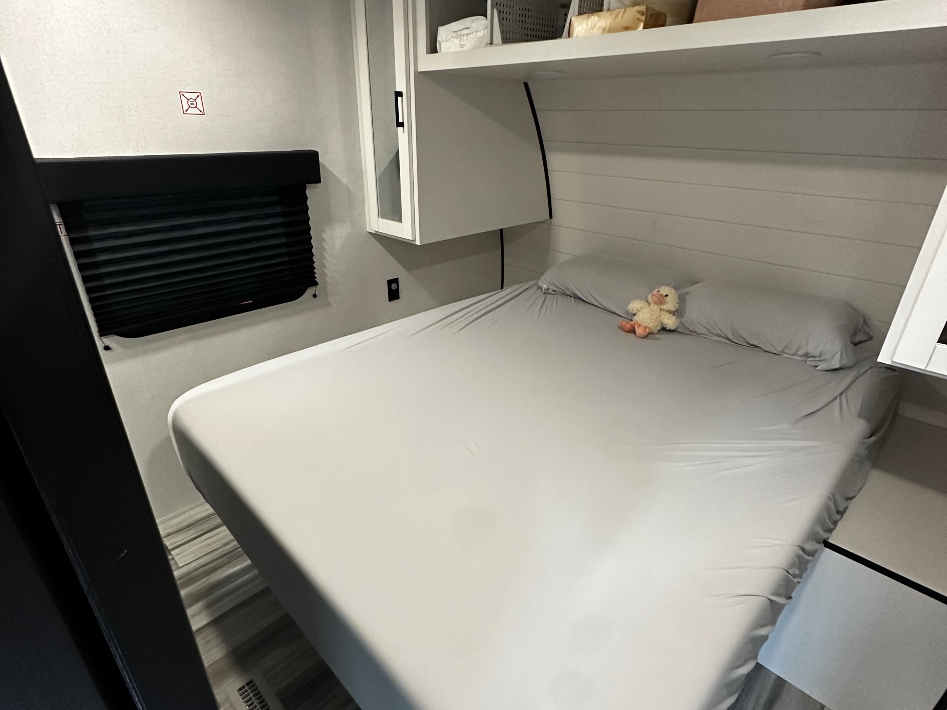 This RV rental features a compact bedroom with a light grey sheeted bed, a plush duck, overhead storage, and a window blind.. Jayco Jay Flight SLX 2026