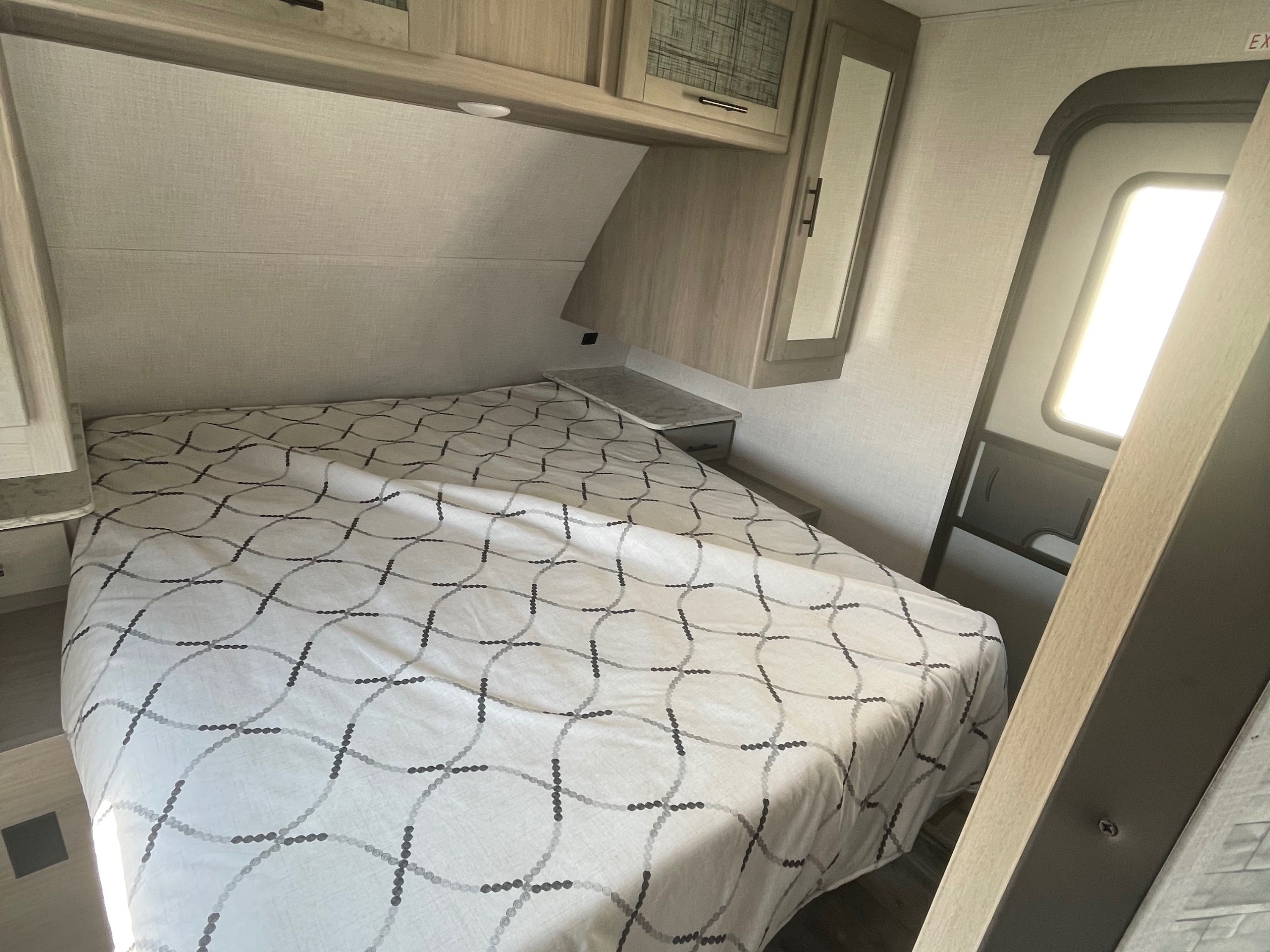 This RV rental's bedroom features a bed with patterned cover, light wood cabinets, and bright lighting from the nearby exit.. Dutchmen Coleman Light 2022