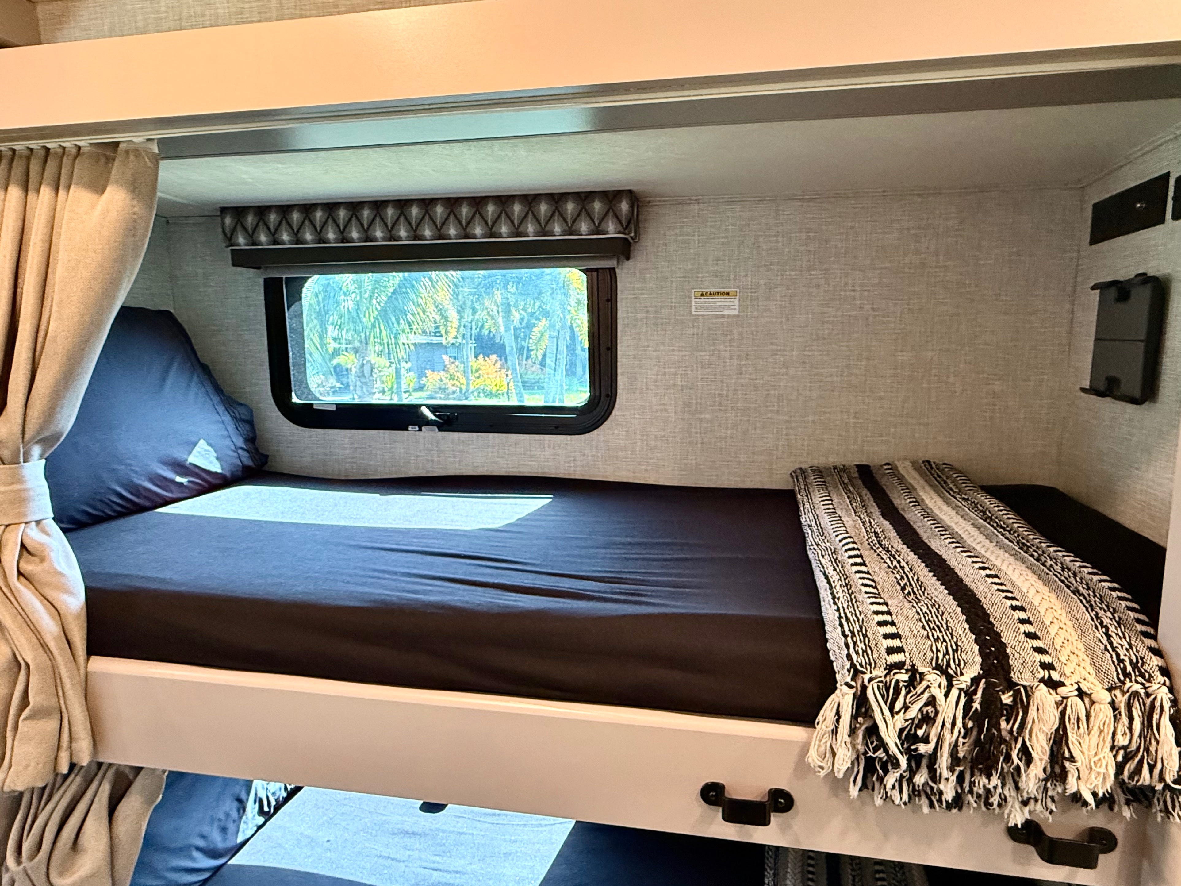 Cozy bunk bed with window view, fitted sheet, and blanket in an RV rental. A compact sleeping area ideal for camping.. Thor Quantum 2021