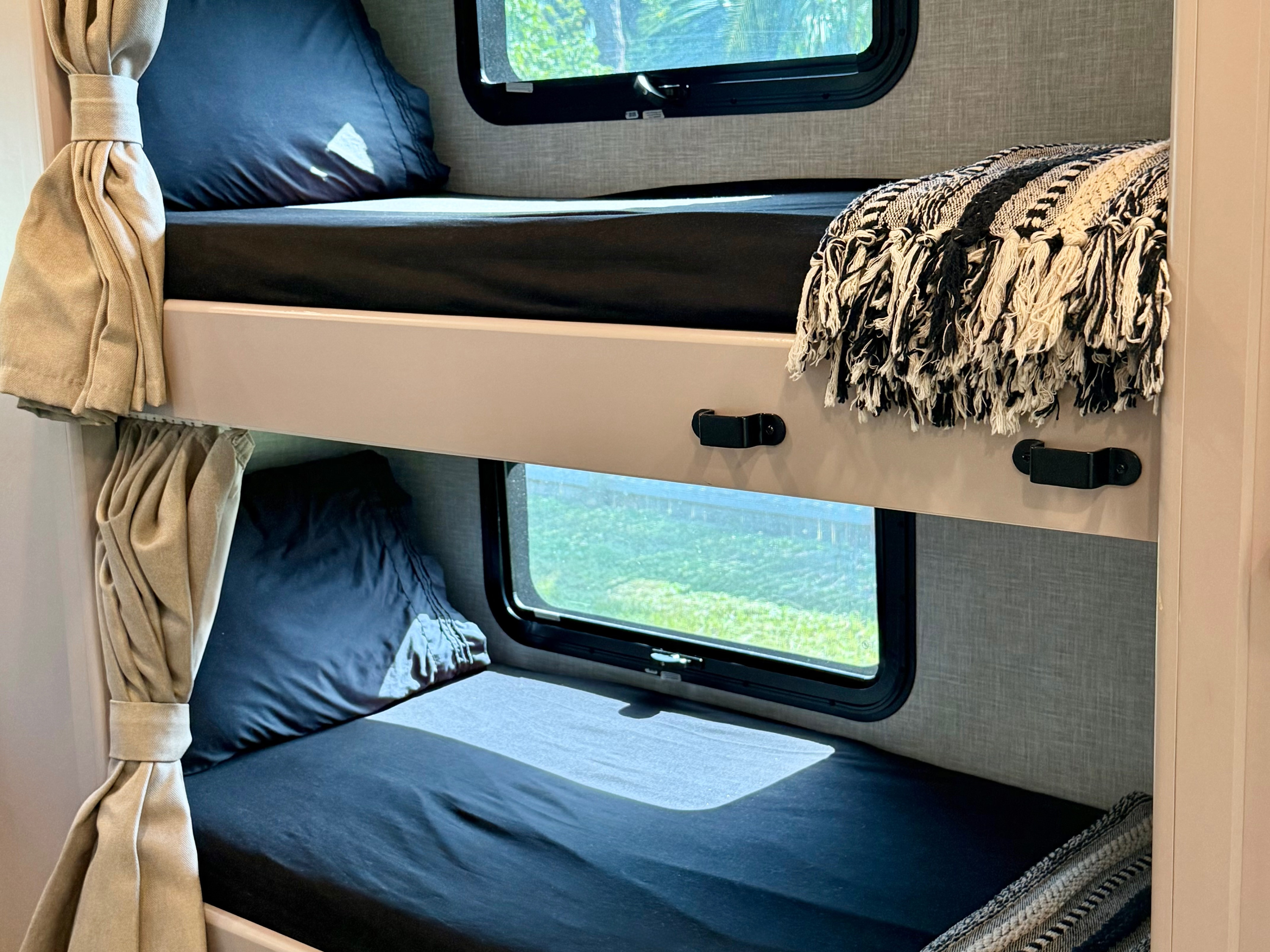 Inside a cozy RV rental: Two bunk beds with dark bedding, tie-back beige curtains, and windows showing outdoor greenery.. Thor Quantum 2021