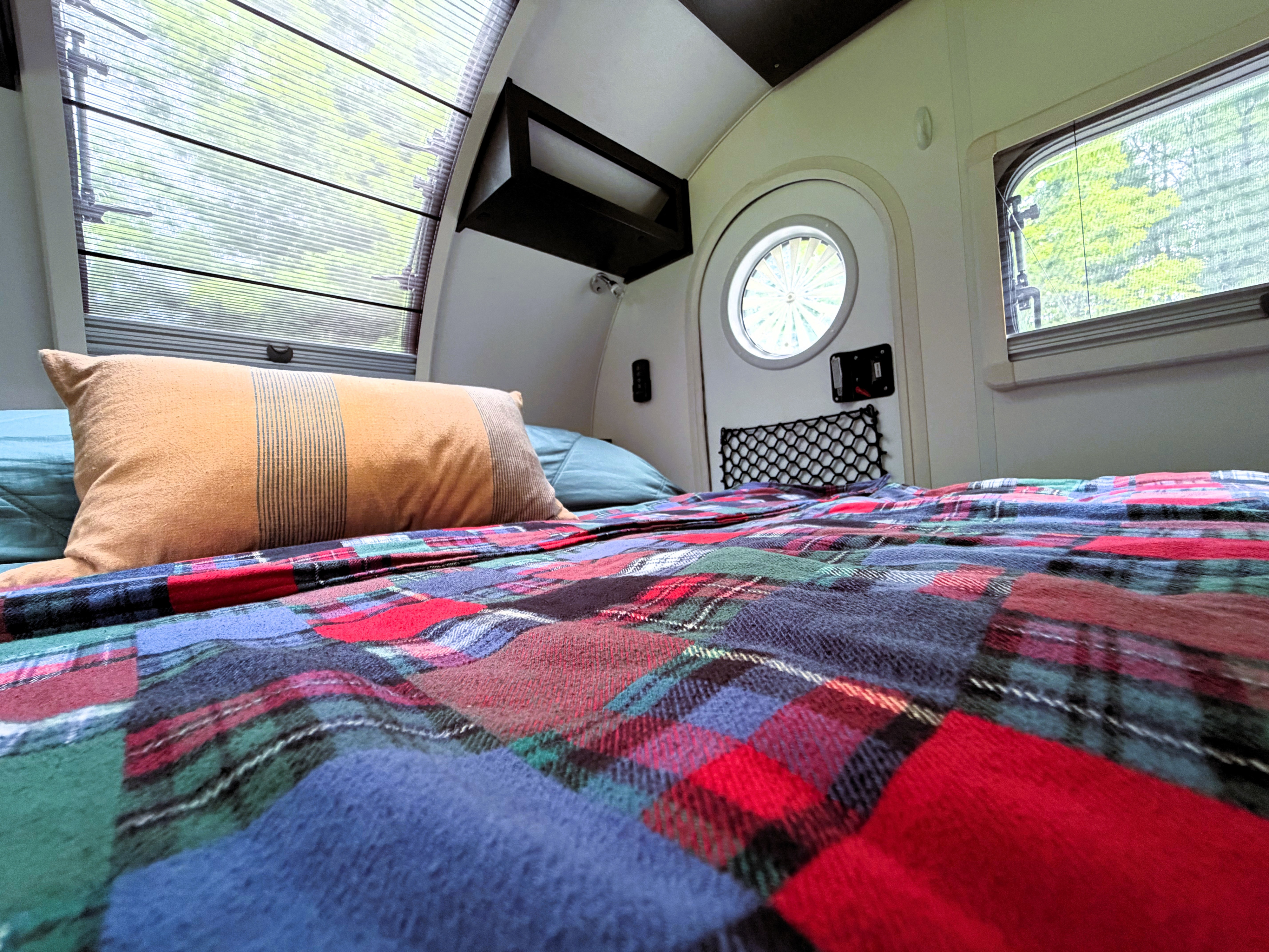 Cozy rv rental bedroom interior, showcasing a bed with a vibrant plaid blanket, accent pillow, and large windows with outdoor views.. nuCamp T@G 2022