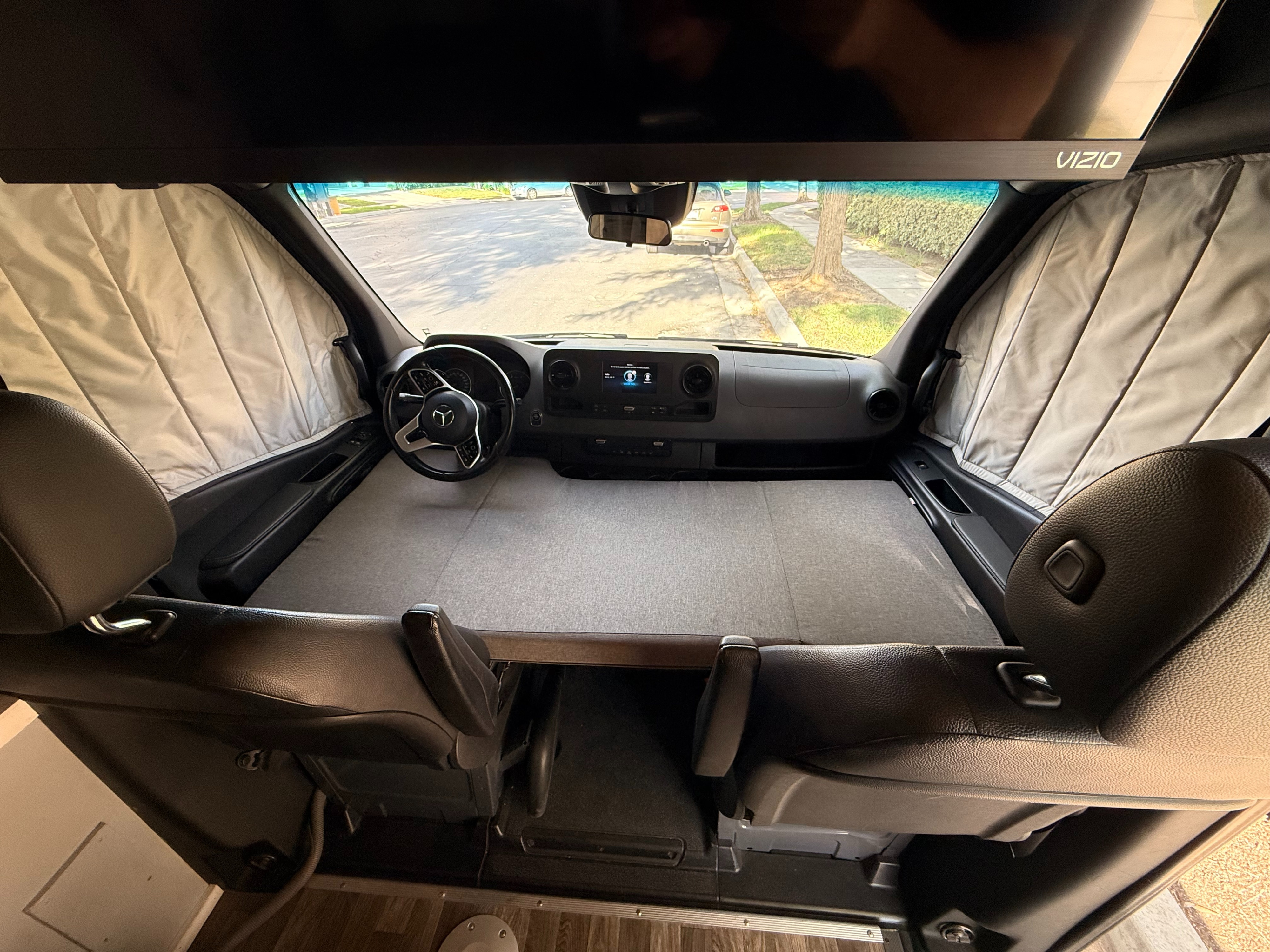 RV rental camper van interior shows front seats, Mercedes dash, bed platform & window shades. Vizio TV above.. Mercedes-Benz Sprinter 2021