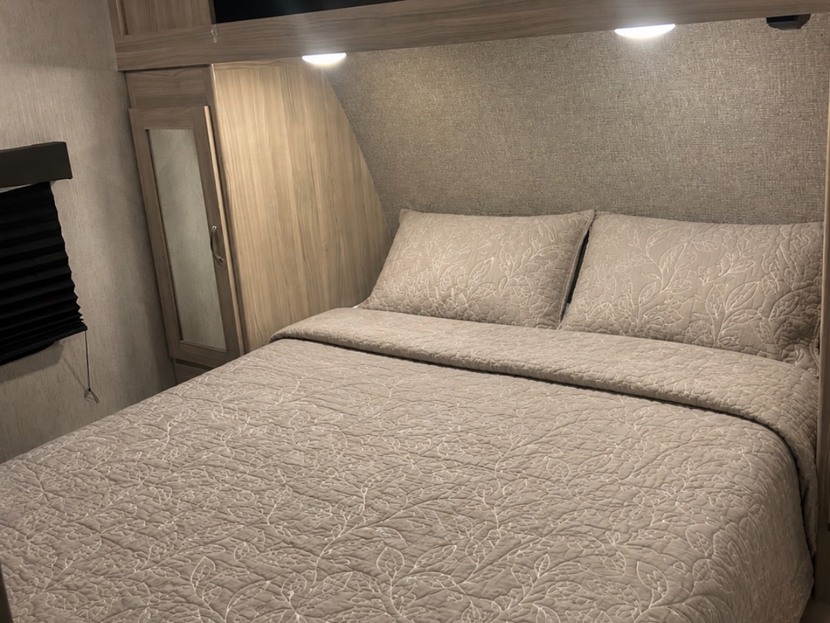 Cozy RV rental bedroom with beige quilted bedding, two pillows, an adjacent wardrobe, and a window blind.. Coachmen Freedom Express 2021