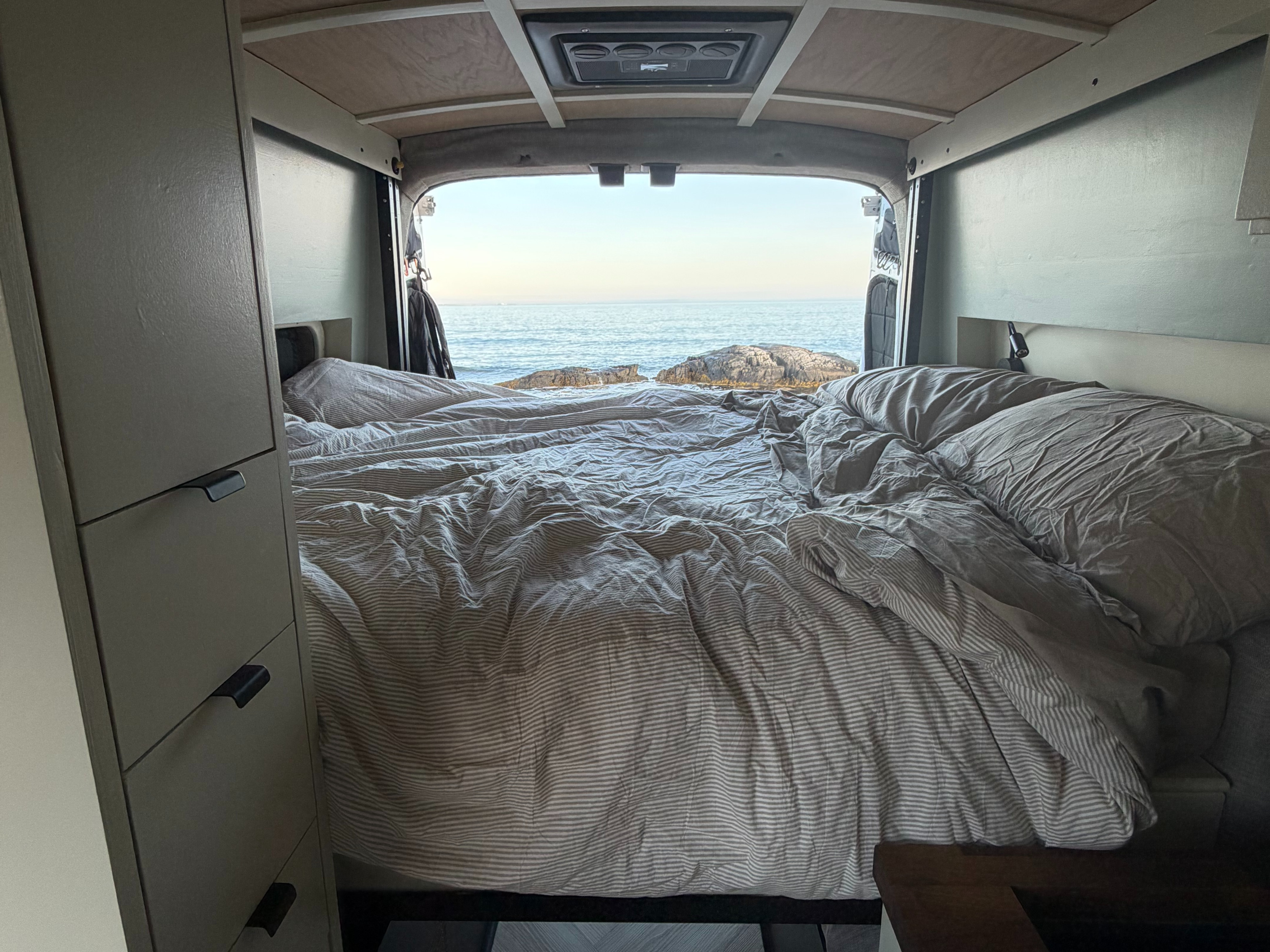 Enjoy a stunning ocean view from an RV rental! Cozy bed with striped bedding inside a camper van, facing a rocky shore and calm sea.. Ford Transit 2020