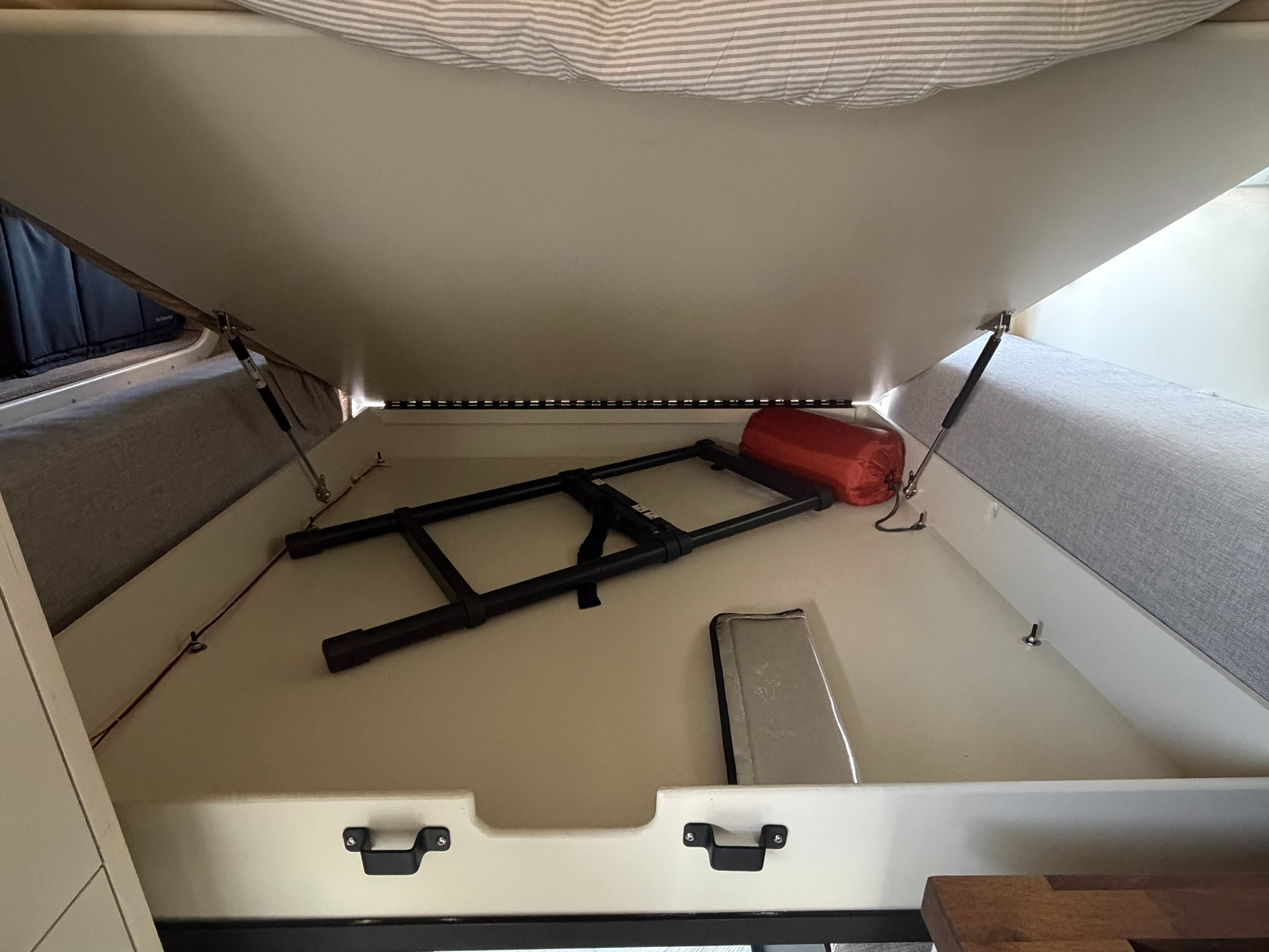 Organized RV rental bed storage compartment, open to show a folding ladder, orange gear bag, and padded items.. Ford Transit 2020