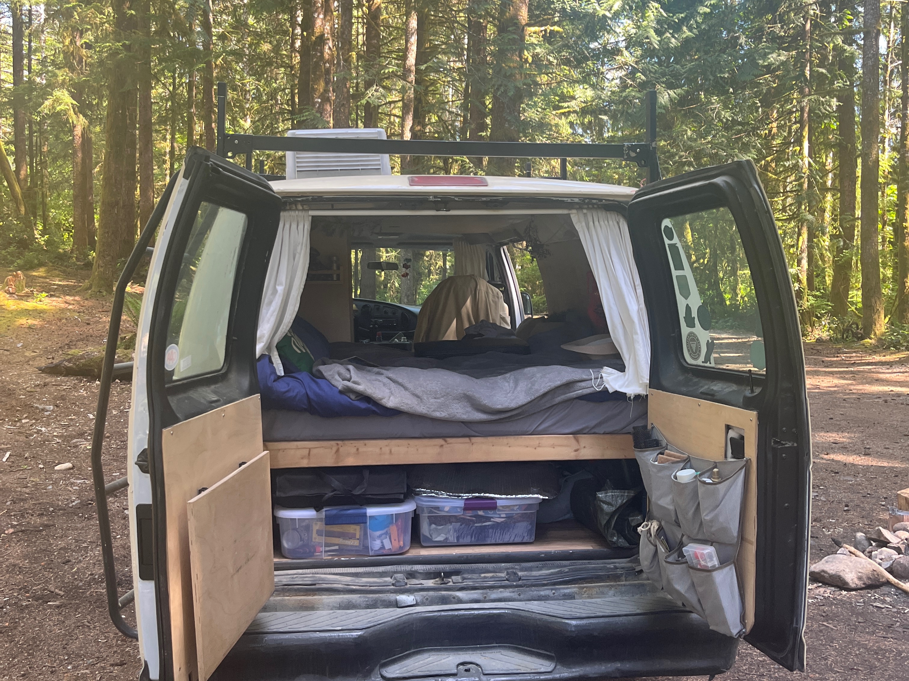 View into the back of an RV rental campervan with open doors, revealing a comfy bed, storage, and gear for forest camping.. Ford E250 Eagle Camper 2005