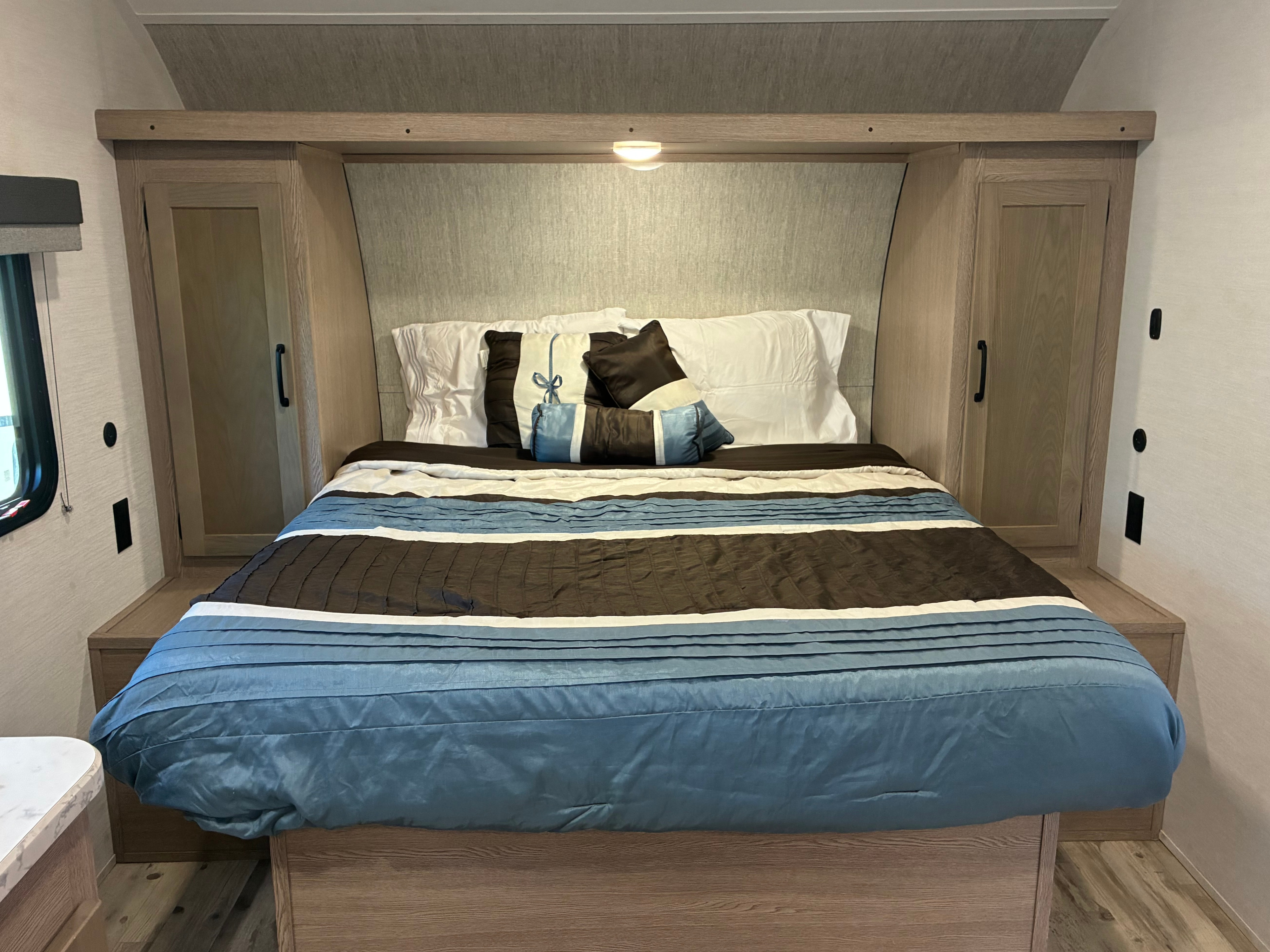 RV rental bedroom: bed with blue, white, brown bedding, decorative pillows. Wooden headboard, cabinets, reading light, window visible.. Gulf Stream Ameri-Lite 2025