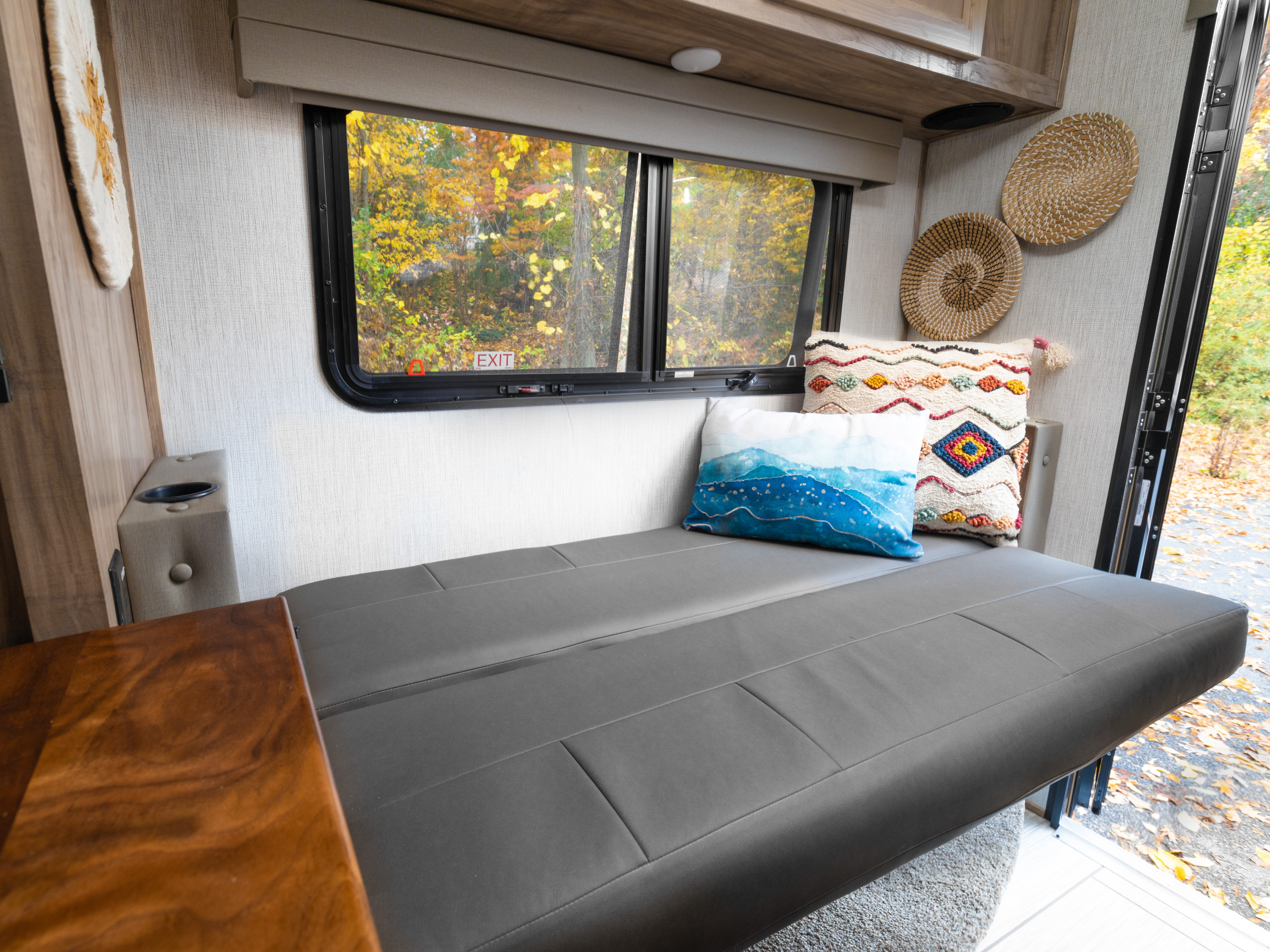 Cozy RV rental interior with a grey fold-down bench-bed, colorful pillows, and a window view of vibrant autumn trees.. Palomino Backpack Edition 2023