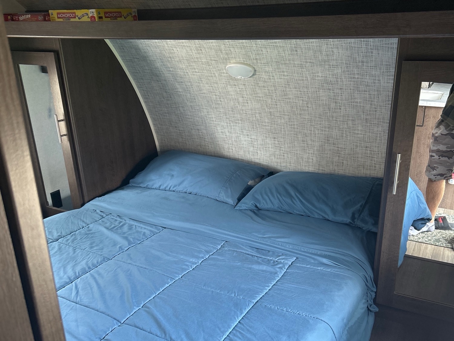 Cozy RV rental bedroom with a blue bed, pillows, and overhead storage with games. Reflects a functional travel space.. Forest River Wildwood X-Lite 2020