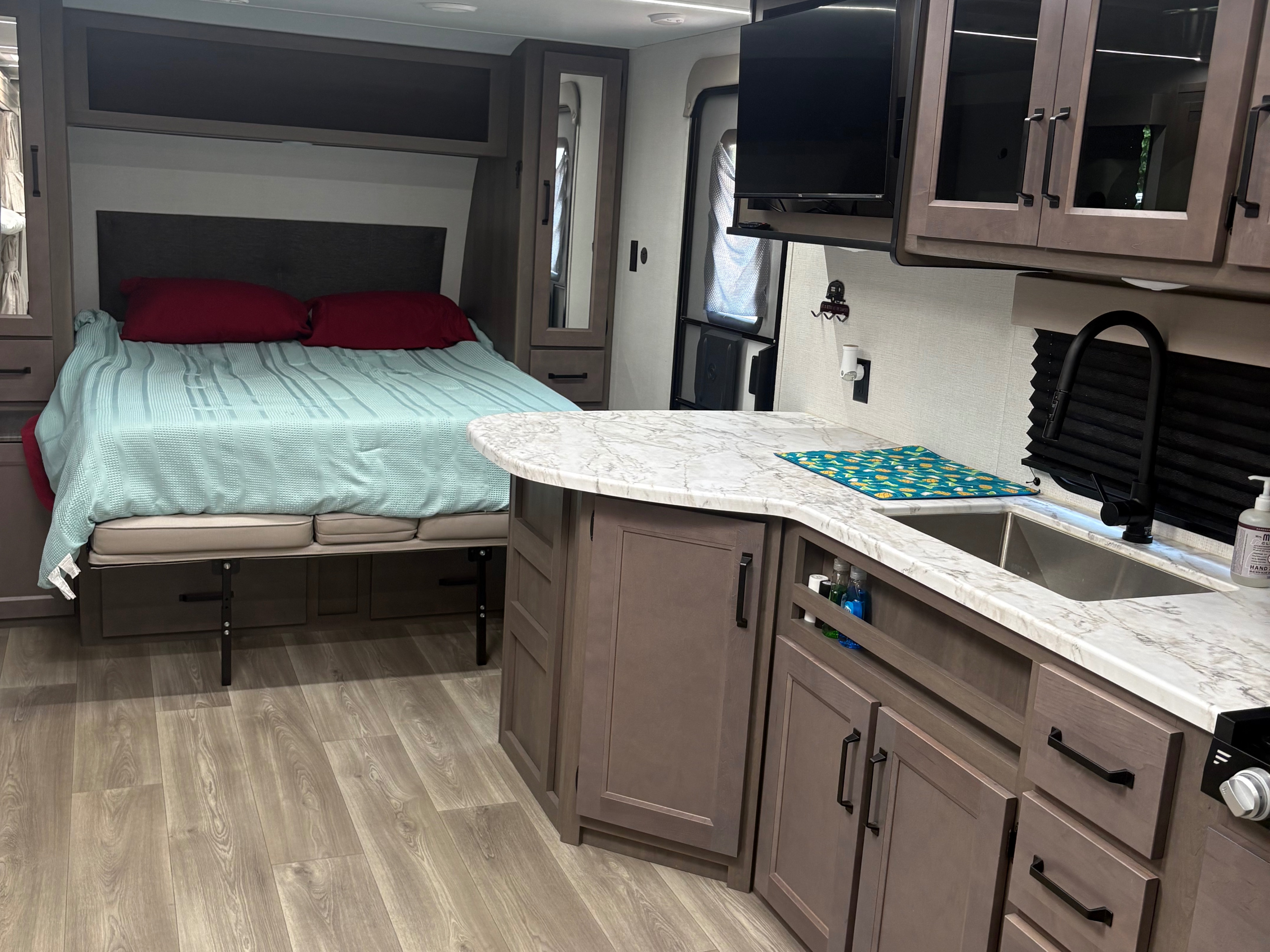 This RV rental interior features a bed with red pillows, kitchen counter with sink, cabinets, and a TV, showcasing a cozy space.. Grand Design Transcend Xplor 2024
