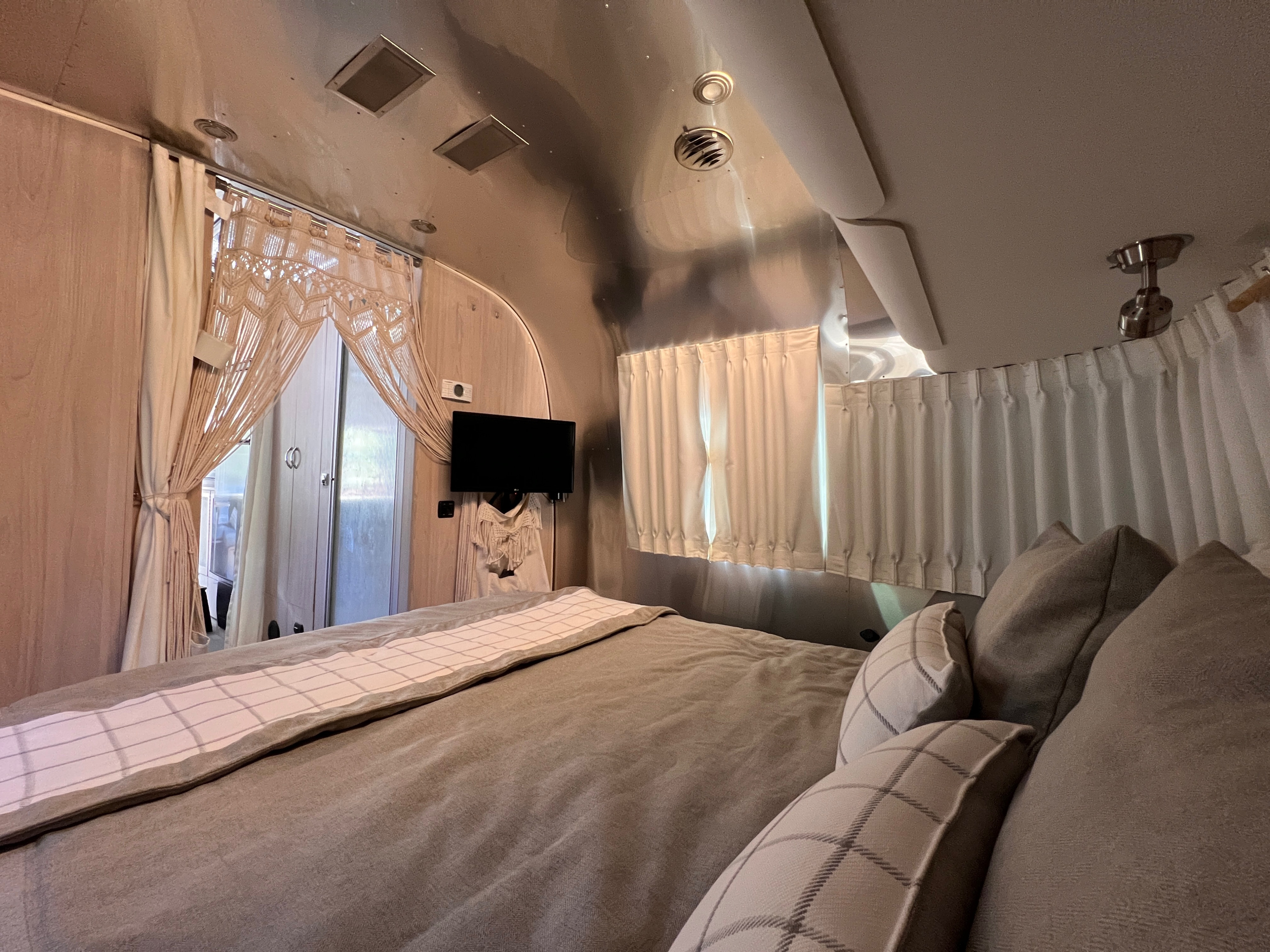 Comfortable rv rental bedroom featuring a large bed, plush pillows, aluminum walls, a mounted TV, and macrame curtains.. Airstream Globetrotter 2022