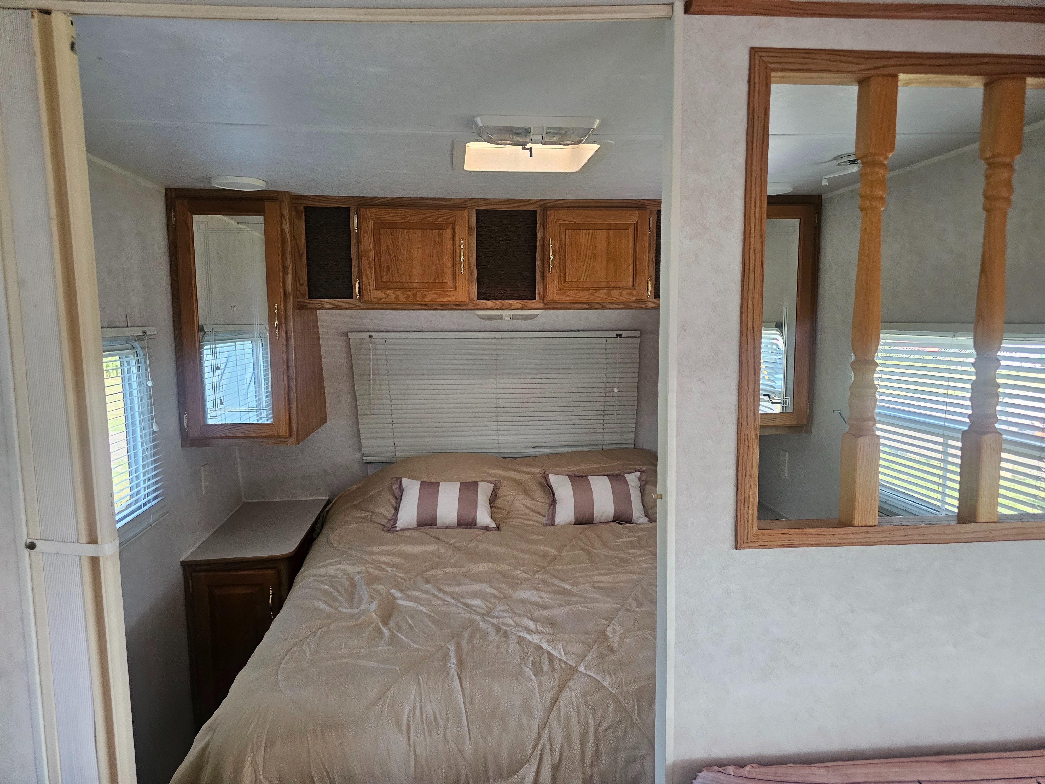 RV rental bedroom: A neatly made bed with striped pillows, wooden overhead cabinets, and window blinds on either side.. Dutchmen Lite 2000