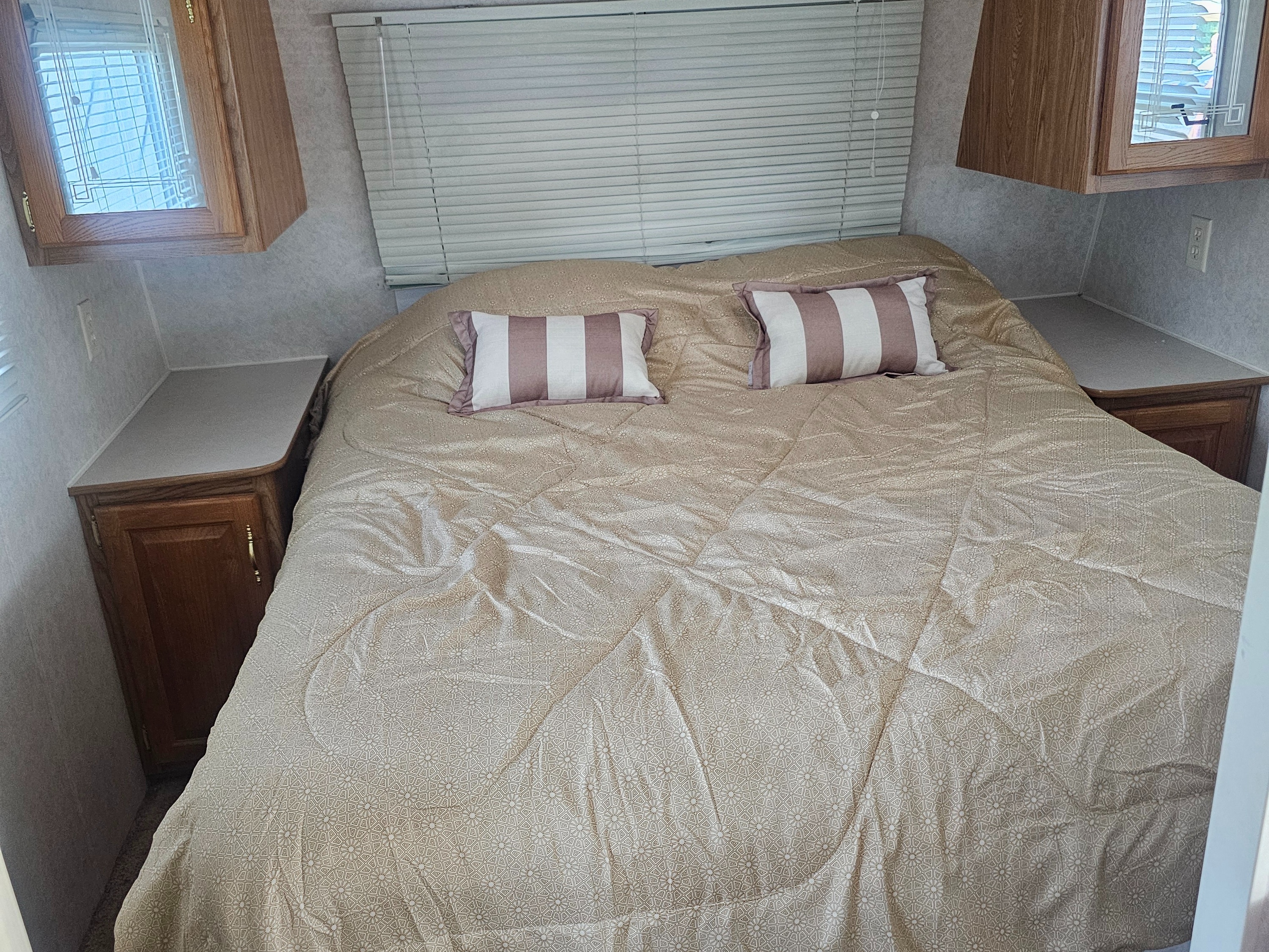 An inviting RV rental bedroom with a bed, two striped pillows, window blinds, and wood cabinetry for a comfortable stay.. Dutchmen Lite 2000