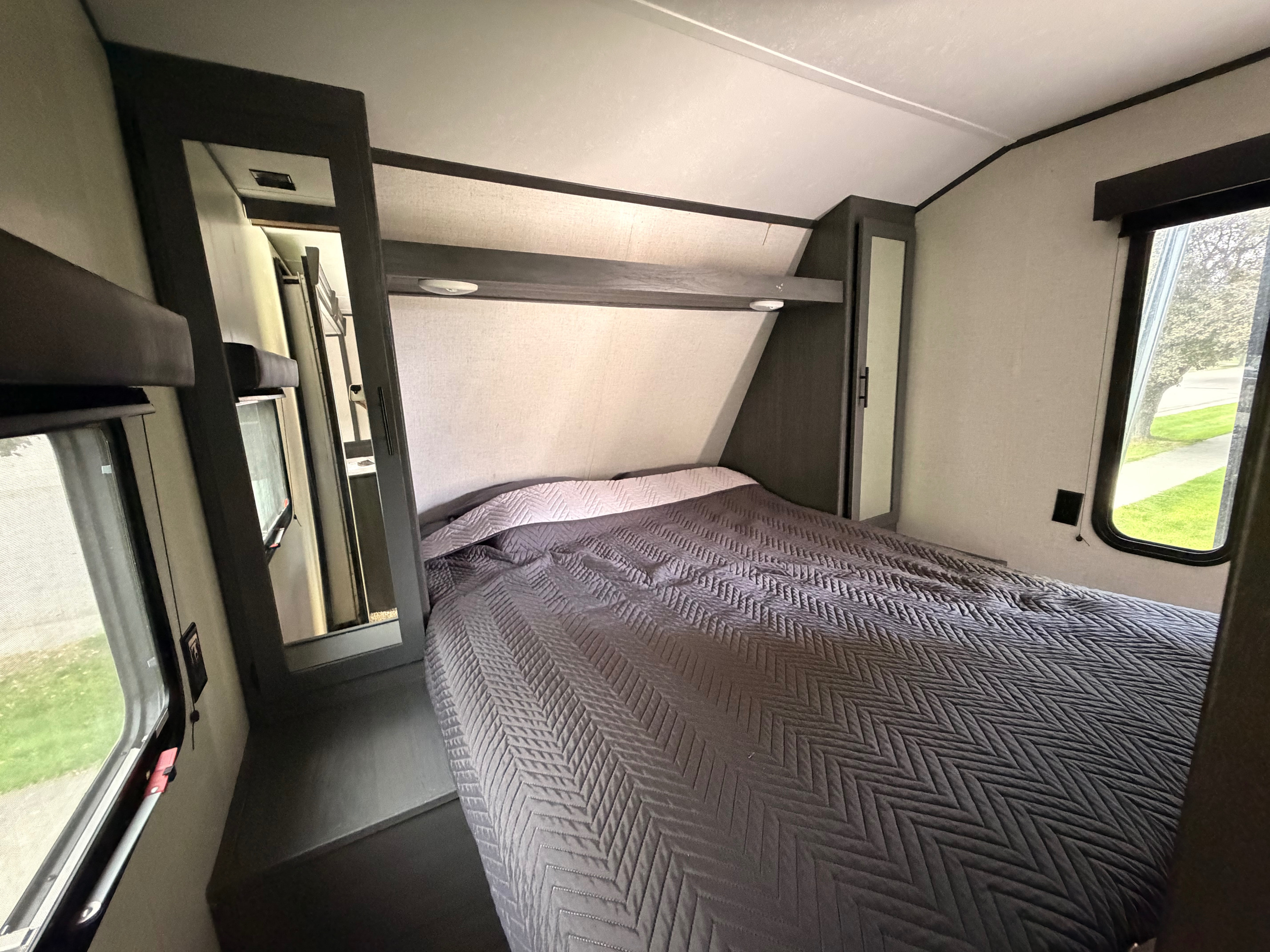 Cozy RV rental bedroom featuring a comfy bed with a dark gray quilted comforter, overhead storage, and two windows.. Aspen Trail Aspen Trail Trailer 2021