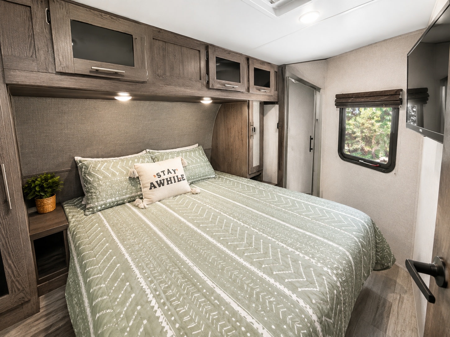 This cozy RV rental bedroom features a comfy bed with green bedding, overhead cabinets, and a window view.. Forest River Surveyor 2018