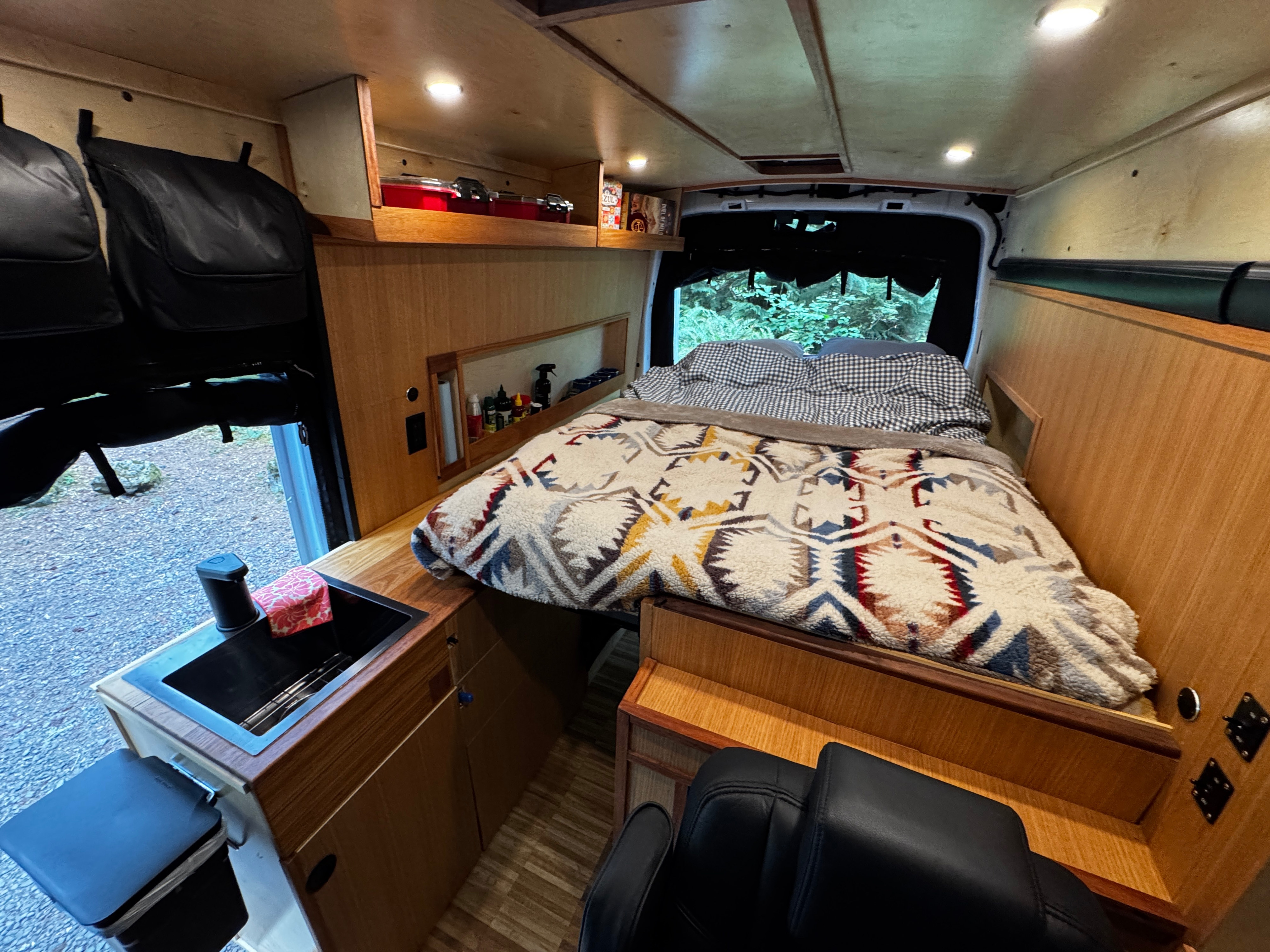 Cozy rv rental interior: a bed with a vibrant blanket, small kitchen sink, and warm wood paneling. Overhead storage and back window view.. Ford Transit 2025