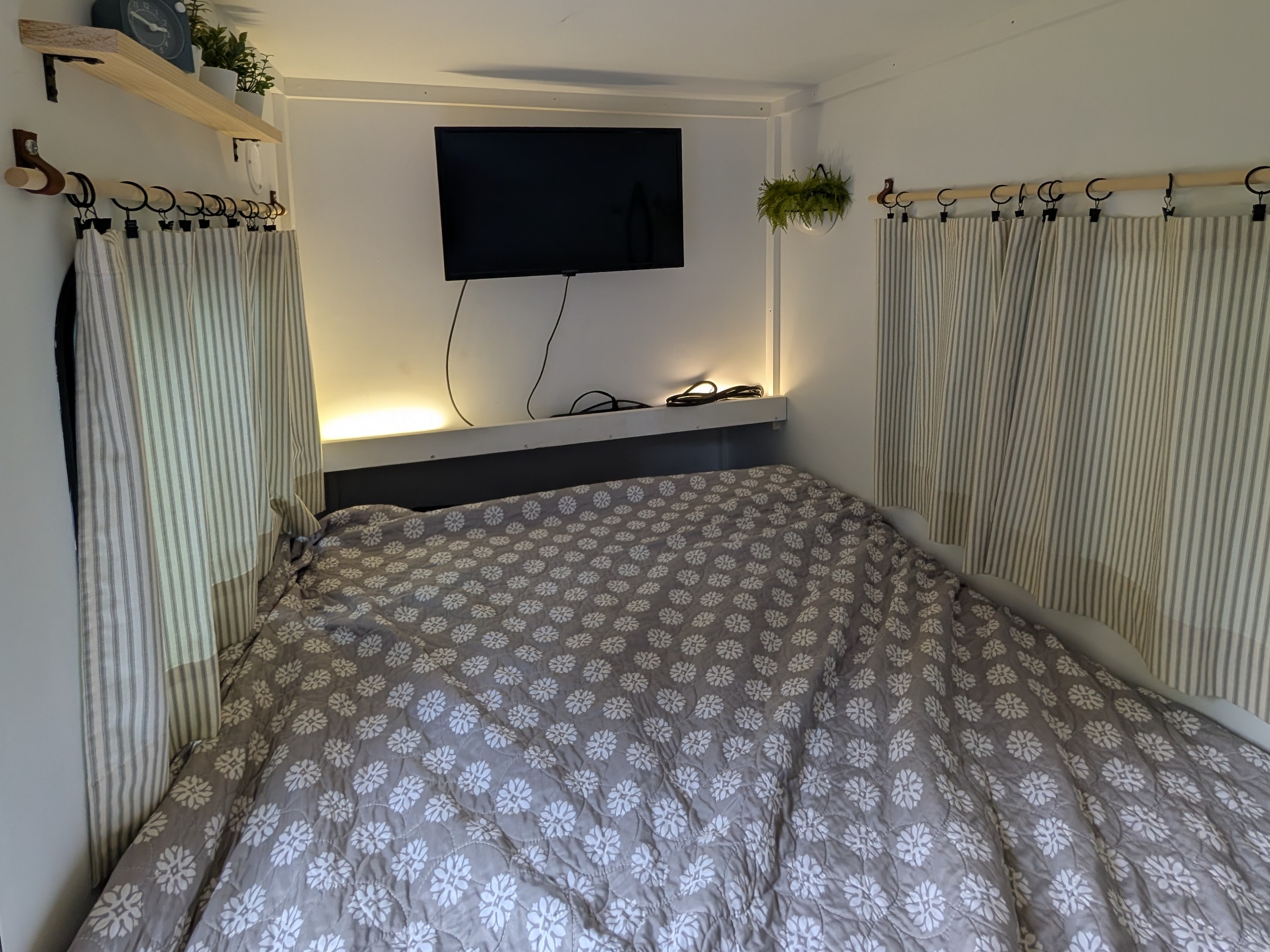 Compact RV rental bedroom featuring a bed with floral quilt, striped curtains, wall-mounted TV, and accent lighting.. MiniTrail Custom 2018