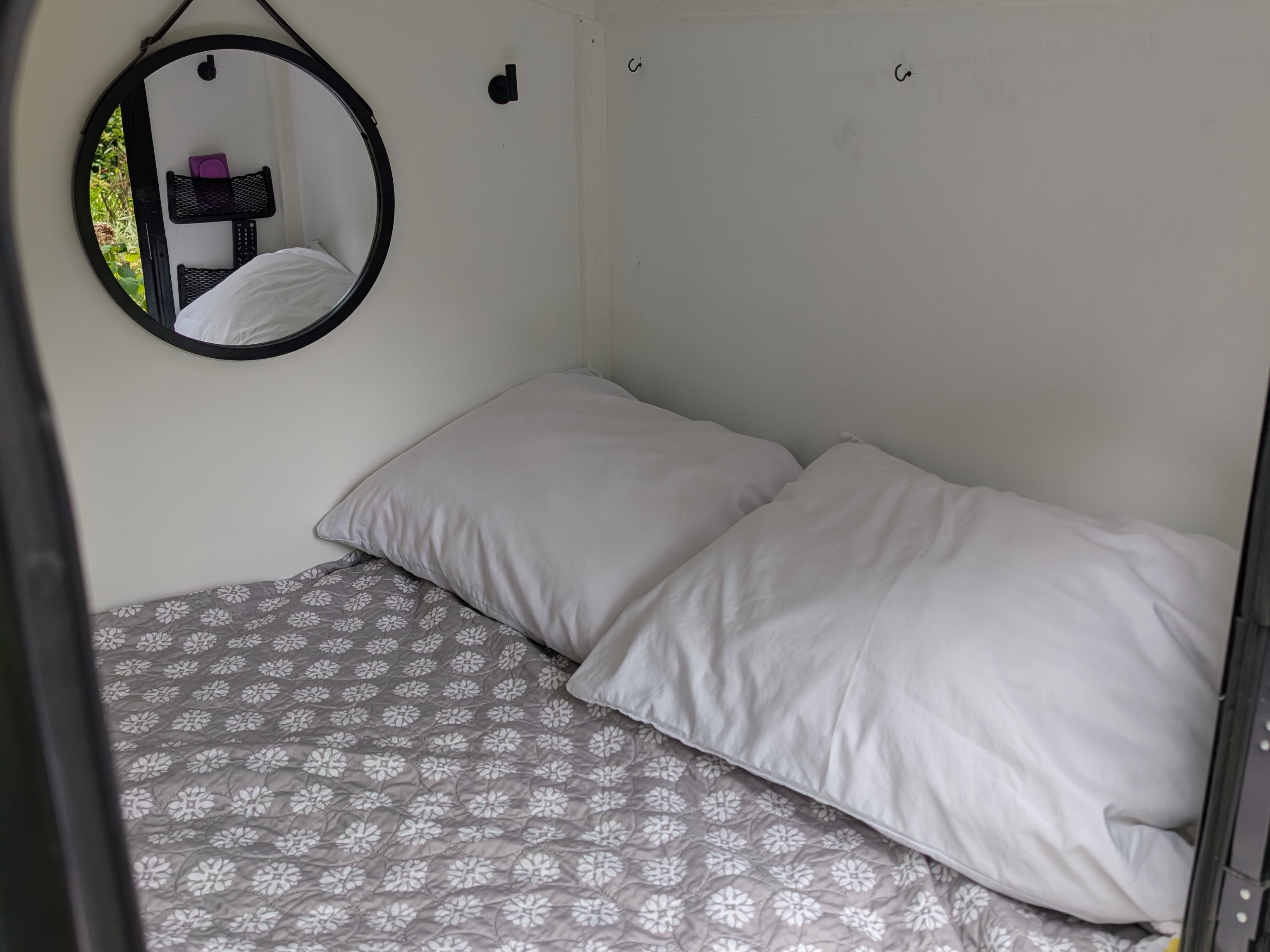 Cozy compact bed in an RV rental with white pillows, a grey floral blanket, and a mirror reflecting greenery.. MiniTrail Custom 2018