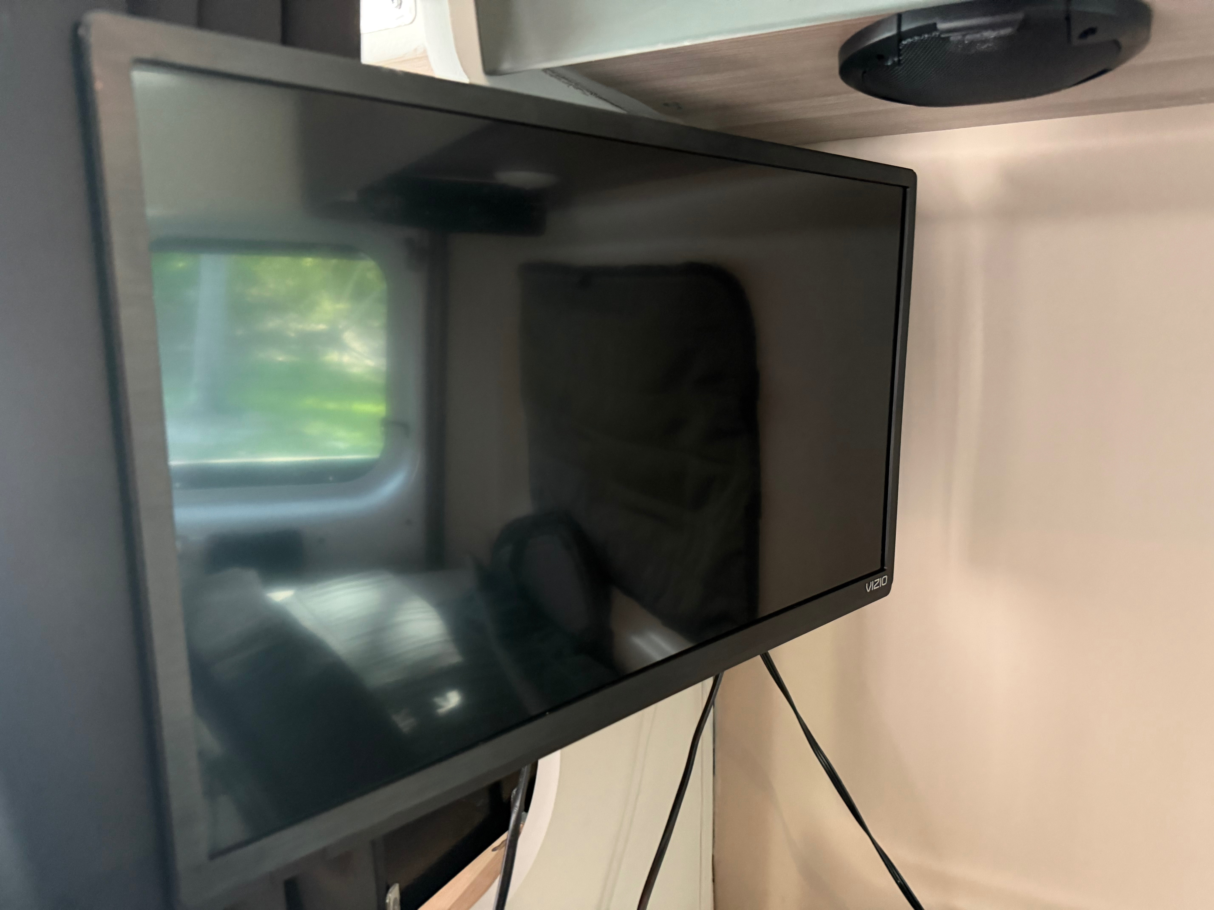 Vizio TV mounted in an RV rental, reflecting a window with green outdoor scenery. A speaker is above, with wires visible.. Dodge 3500shc 2021