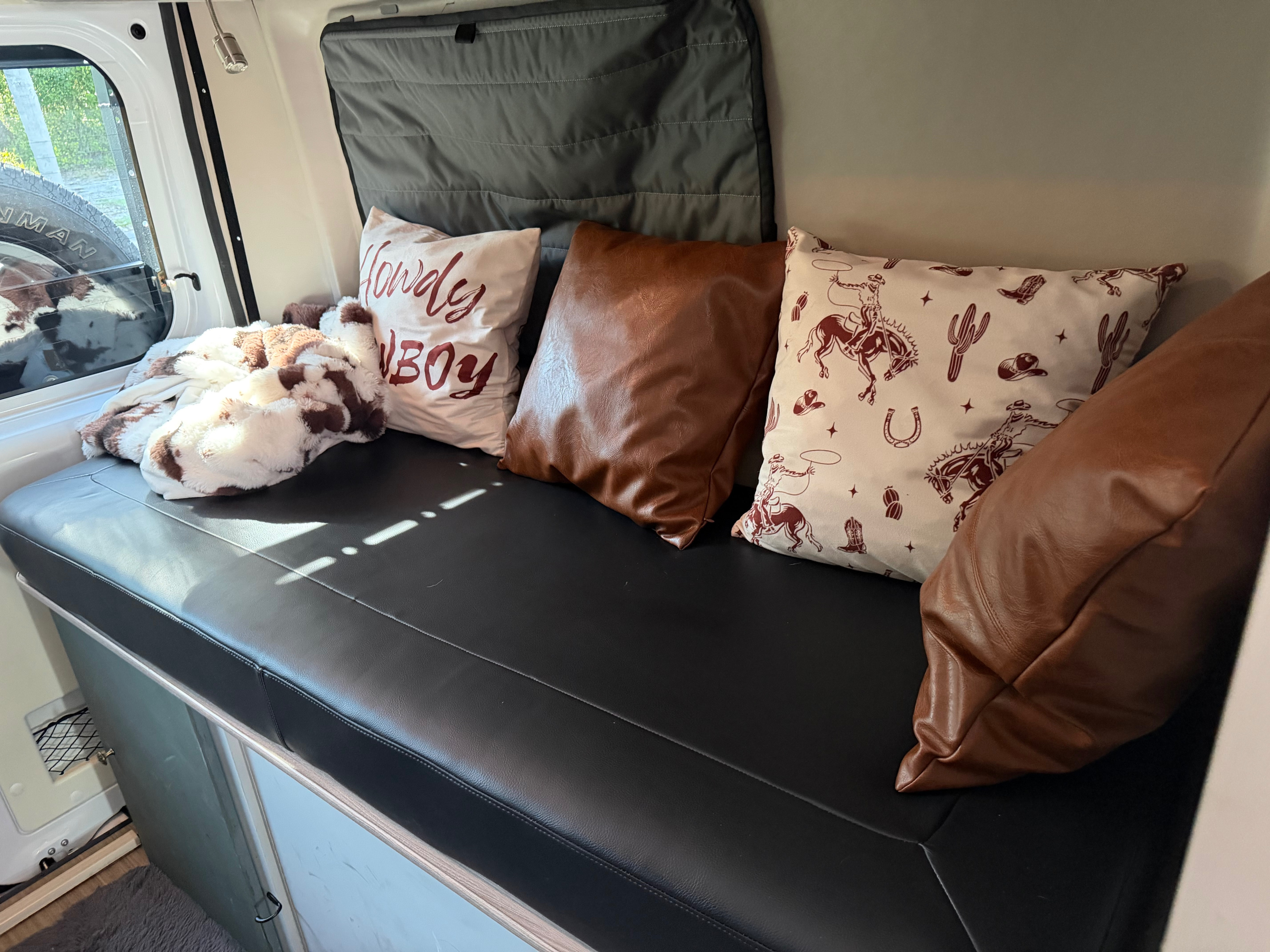 This RV rental features a cozy black bench seat with western-themed pillows and a soft blanket, ideal for relaxing inside the camper.. Dodge 3500shc 2021